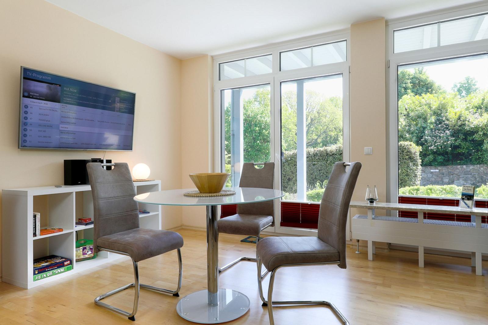 Essbereich mit Glasrundtisch, drei Stühlen und TV an der Wand. Blick auf grünen Garten.