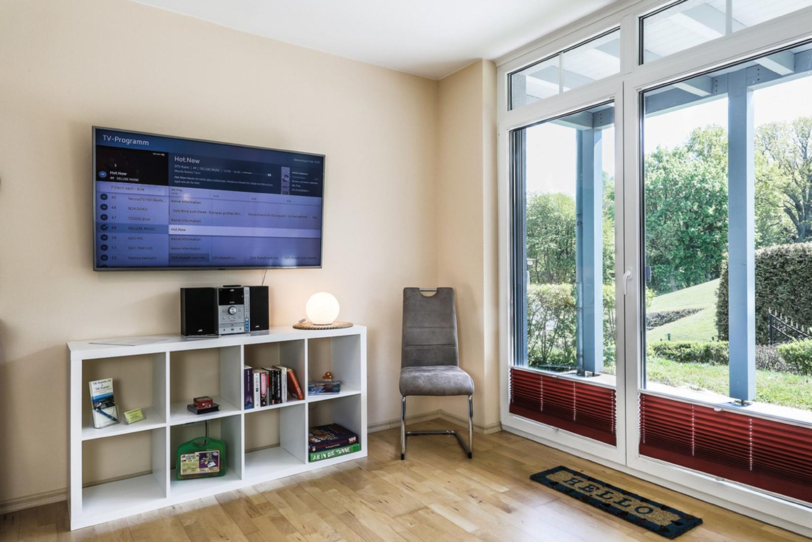 TV-Wand mit Programm, weiße Regale, graue Stuhl und Fenster mit Blick auf Garten.
