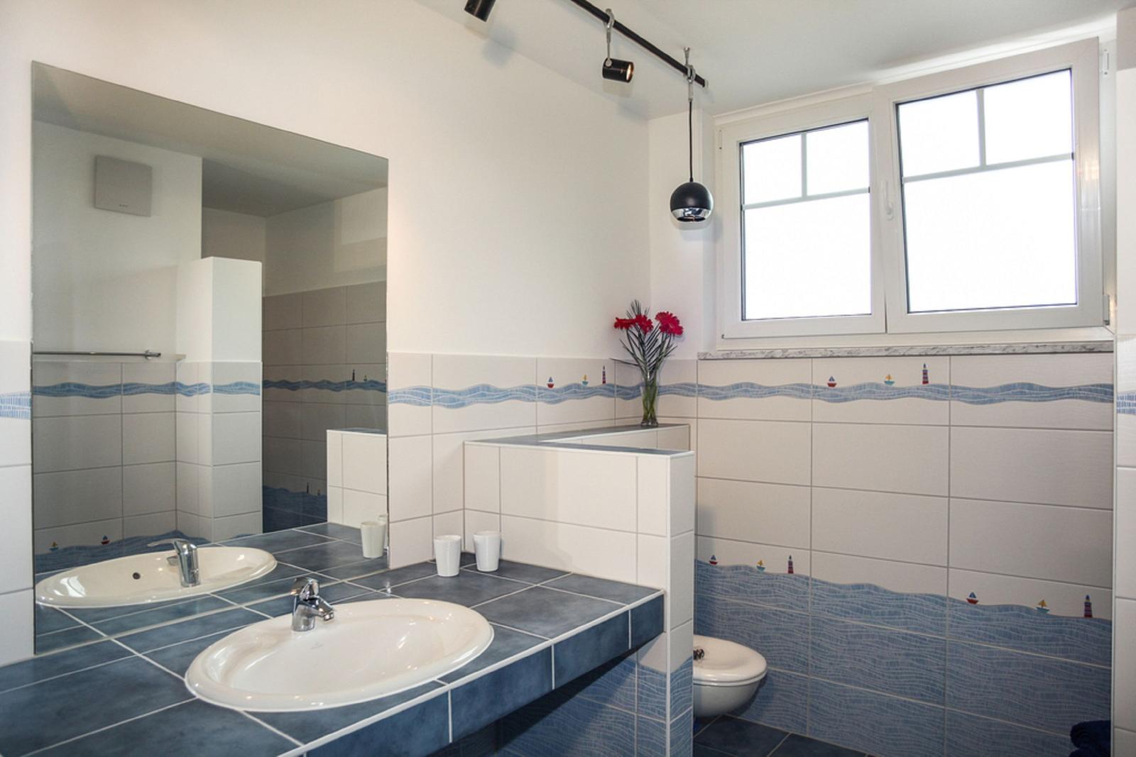 Badezimmer mit zwei Waschbecken, Spiegelschrank und Fenster. Wandfliesen mit Meeresmotiv.