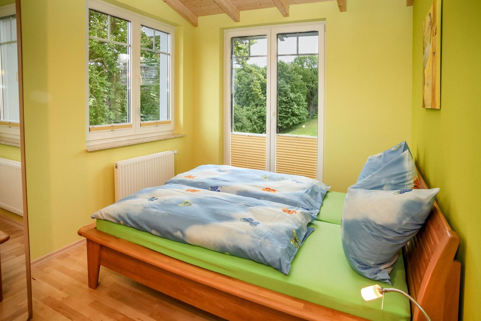 Schlafzimmer mit Holzbett, grünem Matratzenbezug und Fenstern mit Blick auf Grün.