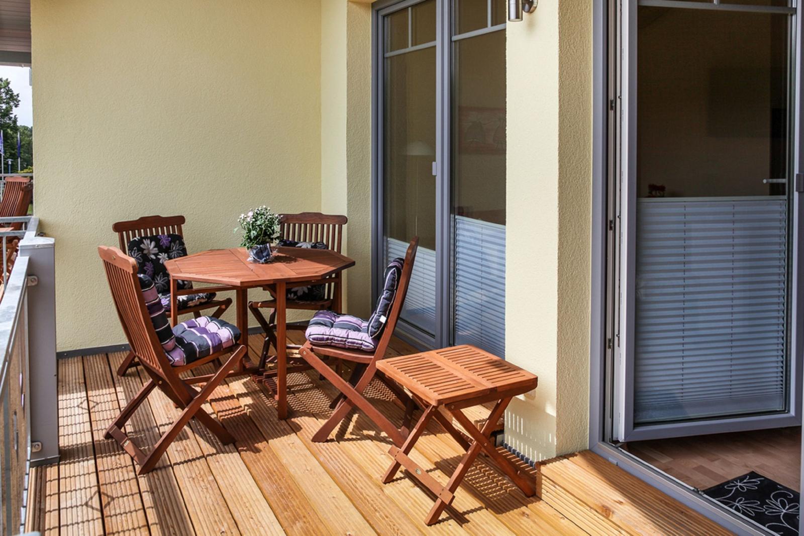 Terrasse mit Holztisch und vier Stühlen mit Kissen, gläserne Schiebetüren und Holzdielen.
