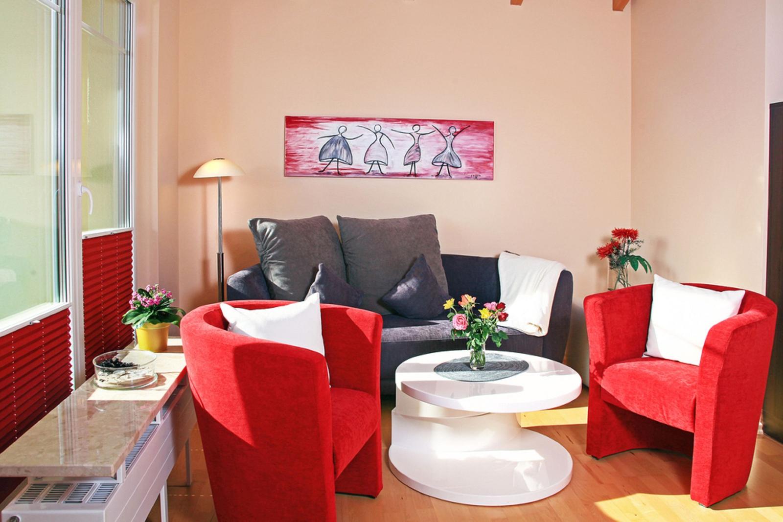 Ein Wohnzimmer mit roten Sesseln, grauem Sofa und runden Tisch. An der Wand hängt ein Bild.