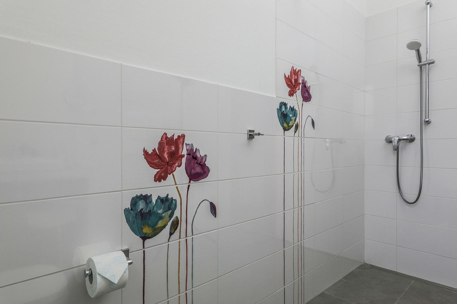 Duschbereich mit weißen Fliesen und bunten Blumenmotiven an der Wand.