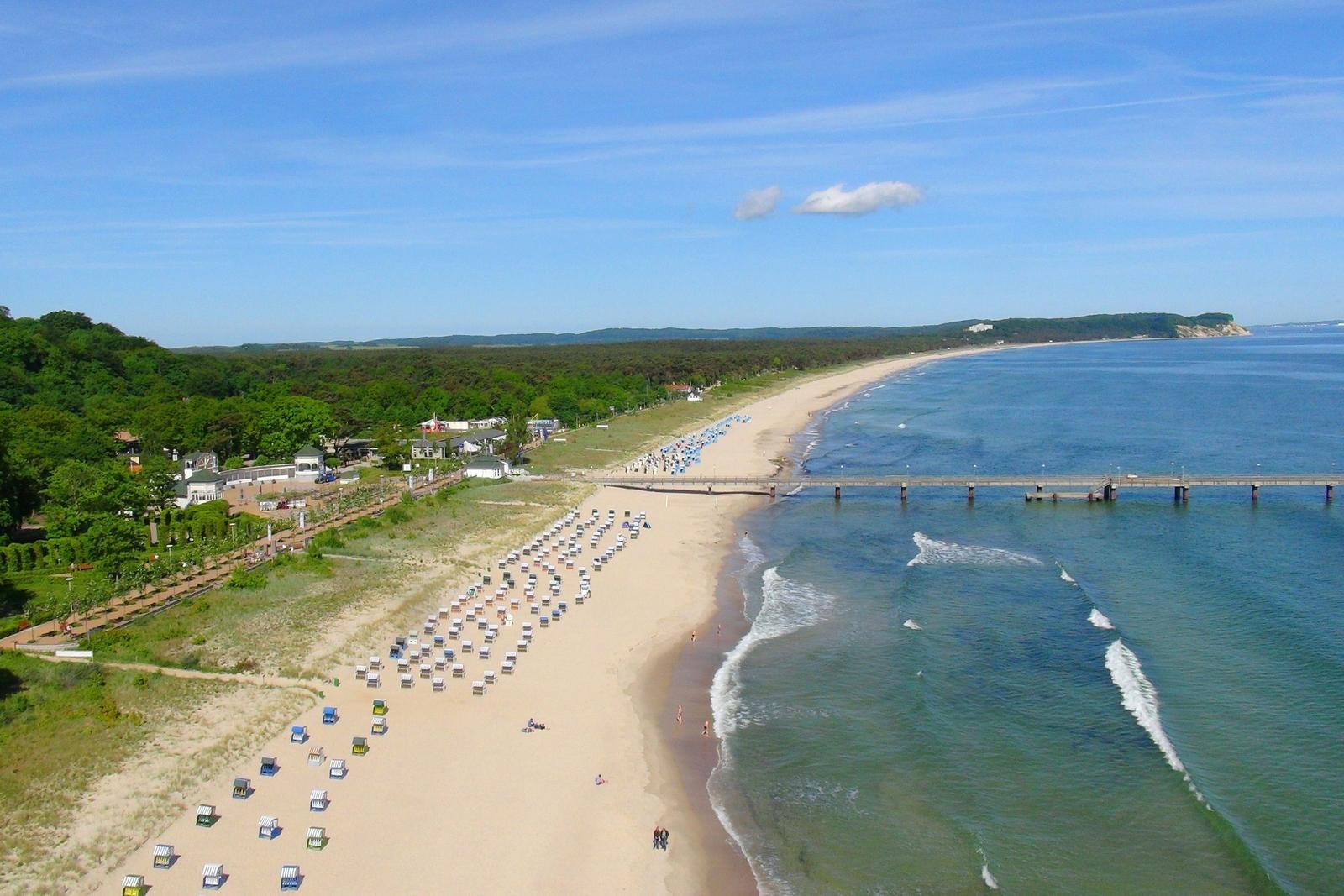 Strand mit Steg, Strandkörben und Wald im Hintergrund unter blauem Himmel.