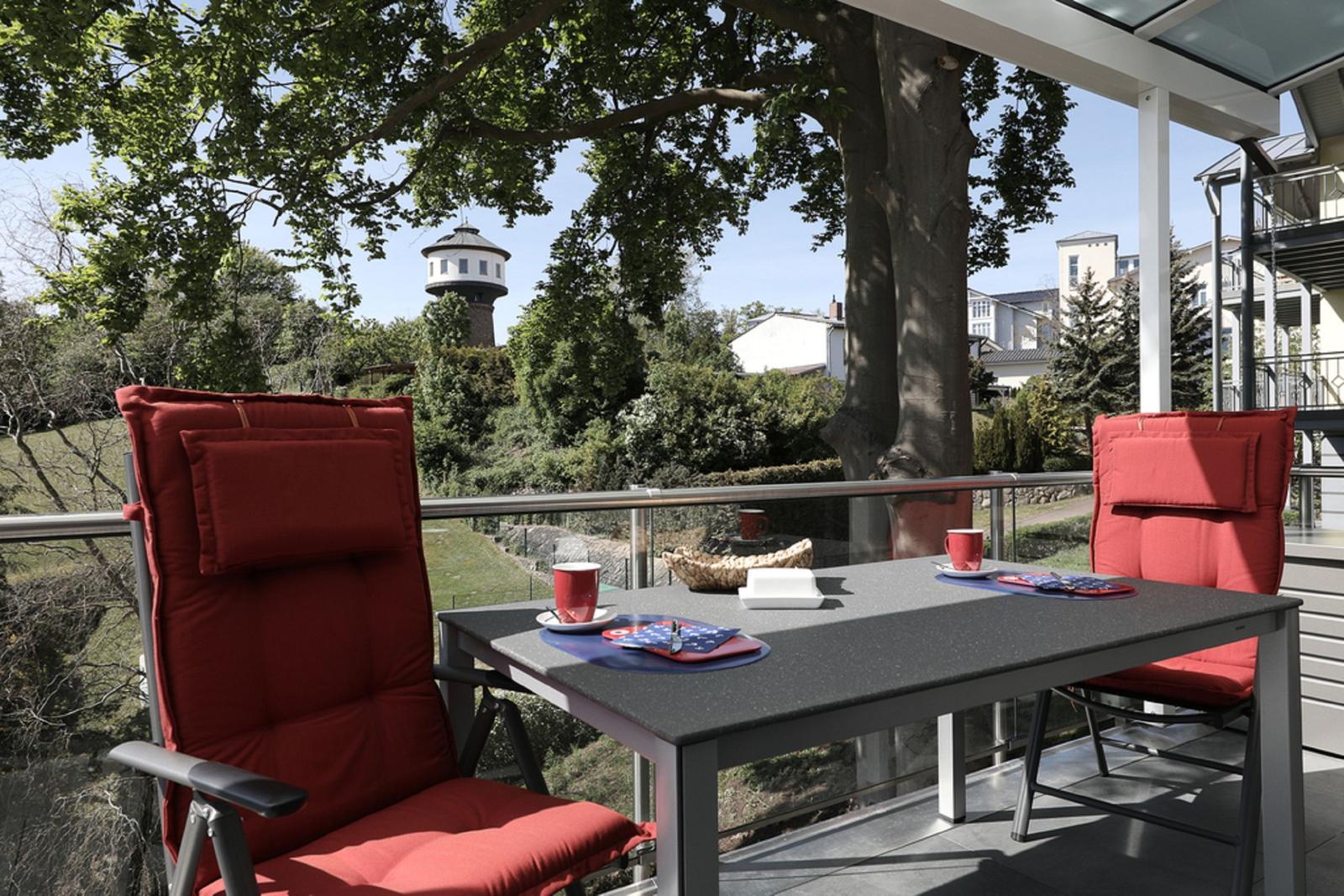 Terrasse mit Tisch und zwei roten Stühlen. Blick auf einen Wasserturm und grüne Bäume.