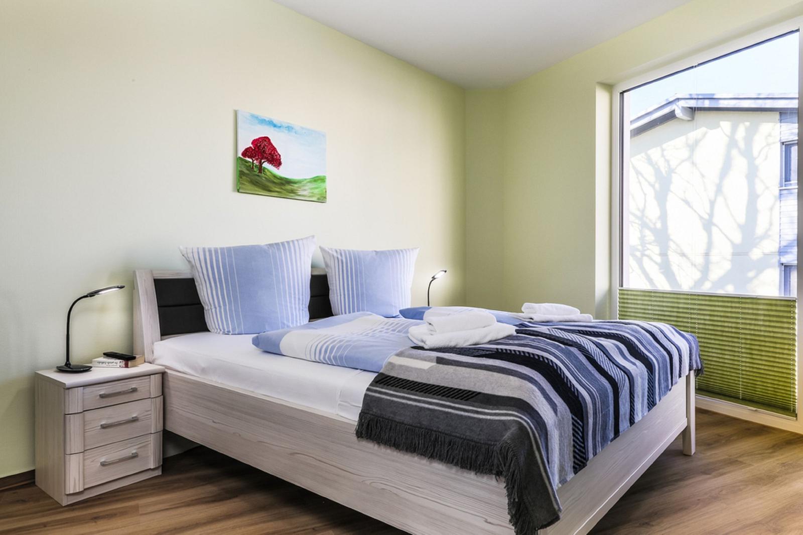 Schlafzimmer mit Bett, Nachttisch und Fenster. Wandbild zeigt einen Baum.