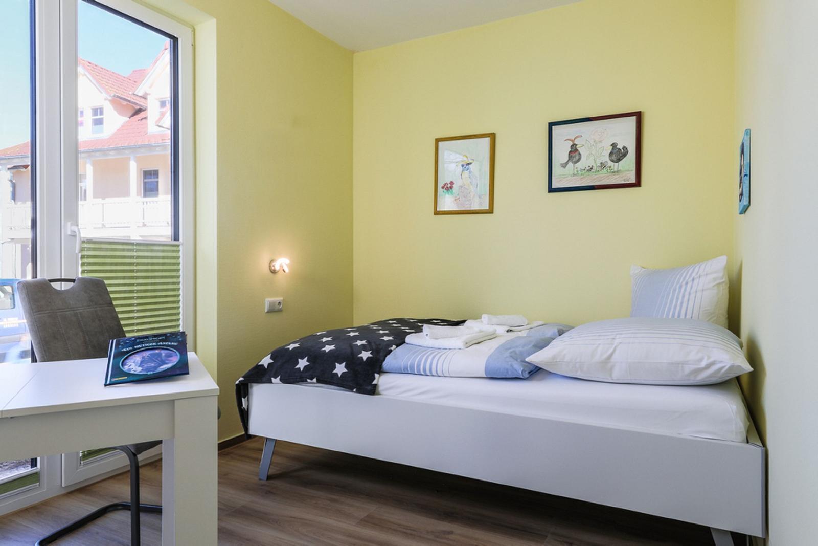 Ein Schlafzimmer mit Bett, Schreibtisch und Fenster. An den Wänden hängen Bilder.