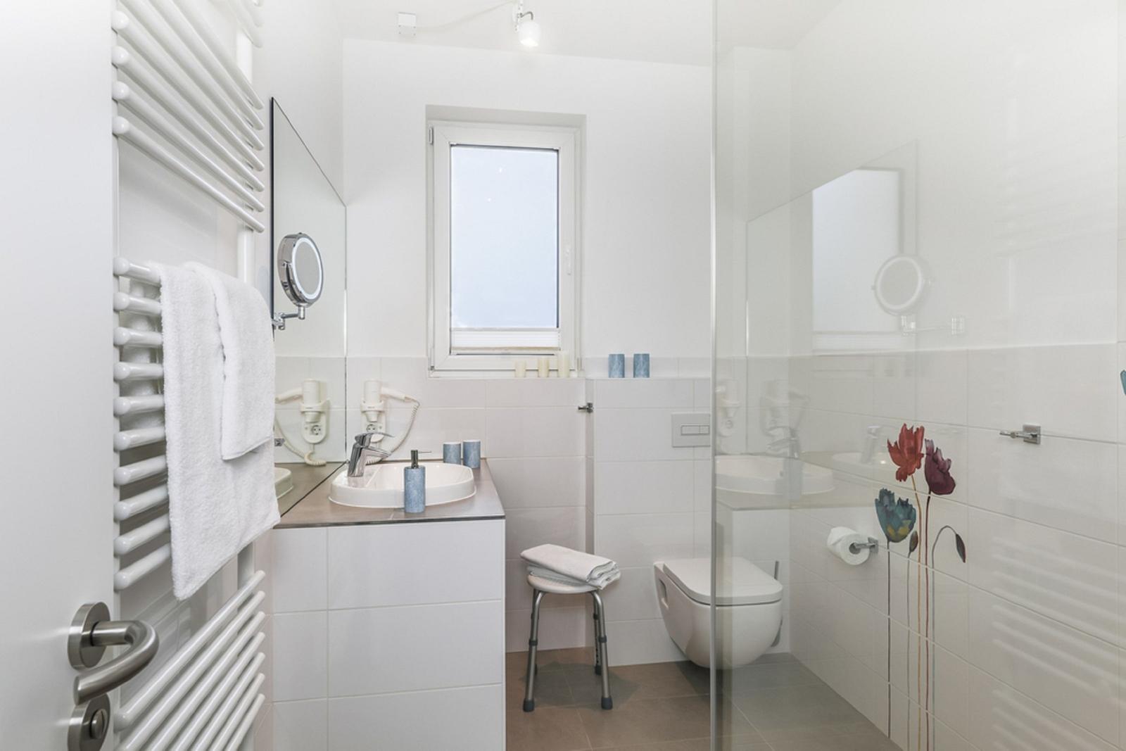 Badezimmer mit Dusche, Waschbecken und Toilette. Weiße Fliesen und Fenster sorgen für viel Licht.