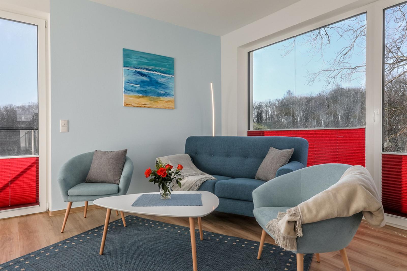 Ein Wohnzimmer mit blauem Sofa, zwei Sesseln und einem weißen Tisch. An den Wänden hängt ein Bild.