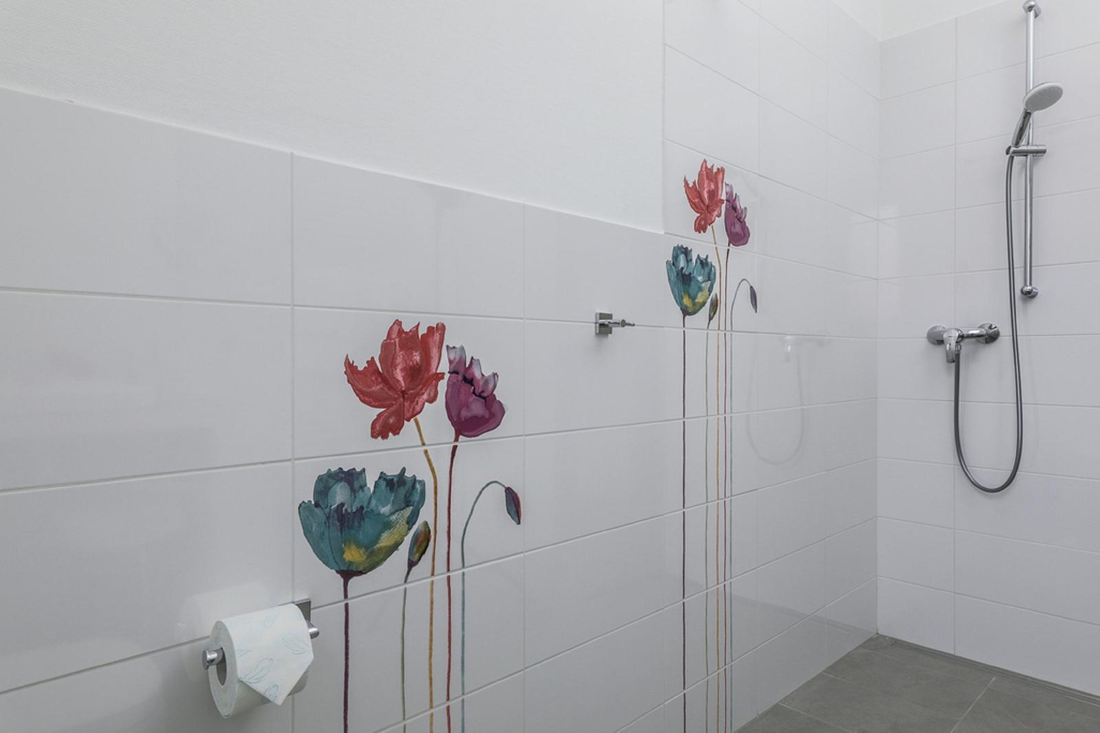 Dusche mit weißen Fliesen und bunten Blumenmotiven an der Wand. Toilettenpapierhalter sichtbar.