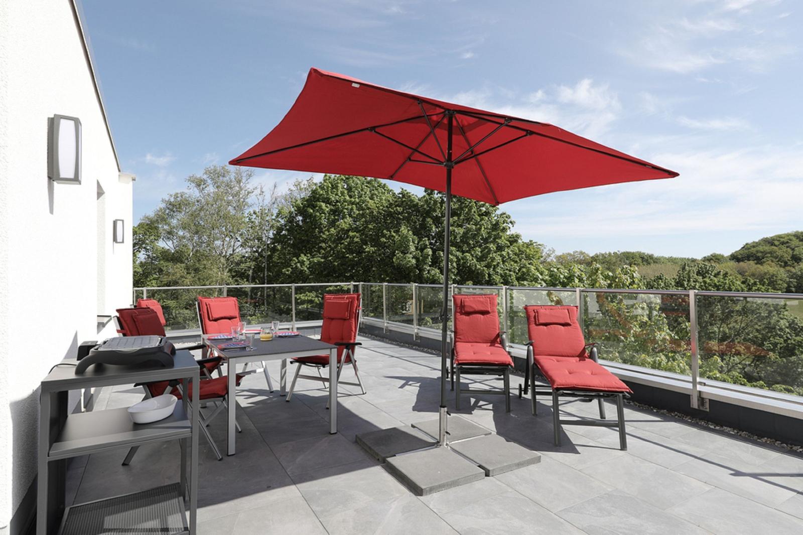Terrasse mit rotem Sonnenschirm und Sitzgelegenheiten, Blick auf grüne Bäume.