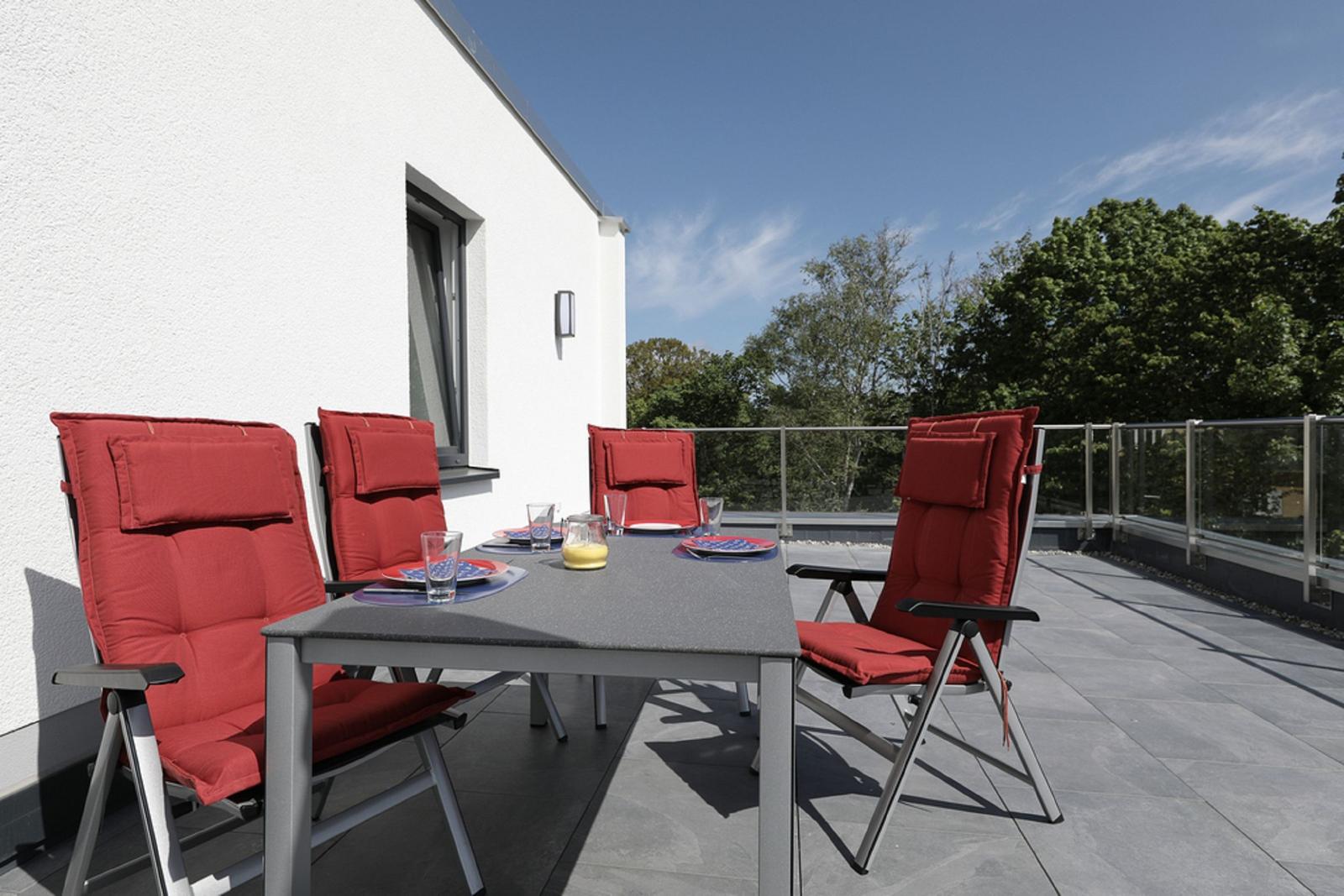 Terrasse mit Tisch und vier Stühlen mit roten Sitzkissen. Hintergrund: weißes Haus und Bäume.