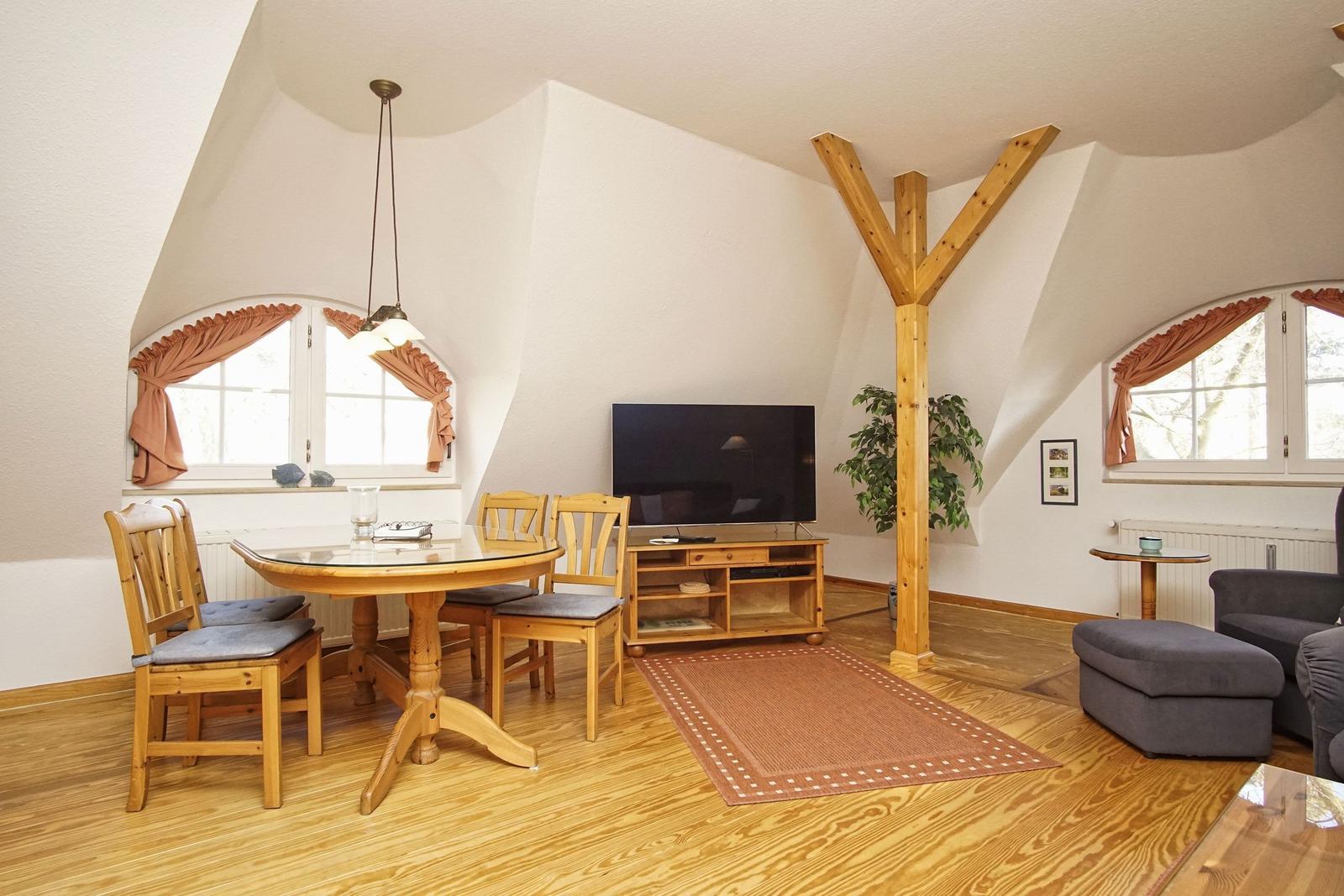 Wohnzimmer mit Essbereich, Fernseher und Holzfußboden. Zwei Fenster mit Vorhängen.