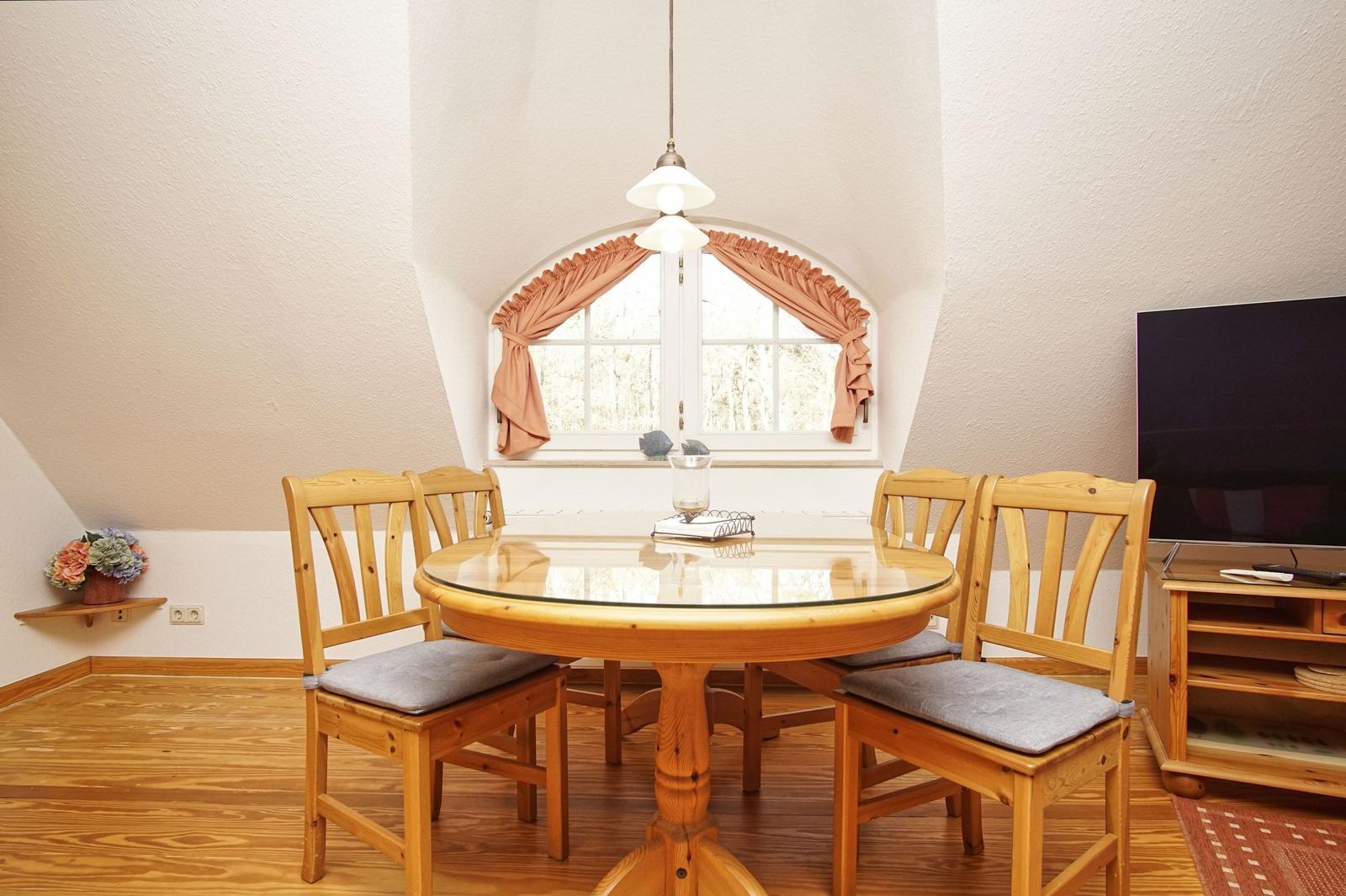 Esszimmer mit Holztisch, vier Stühlen und Bogenfenster. Fernseher und Blumen sind sichtbar.