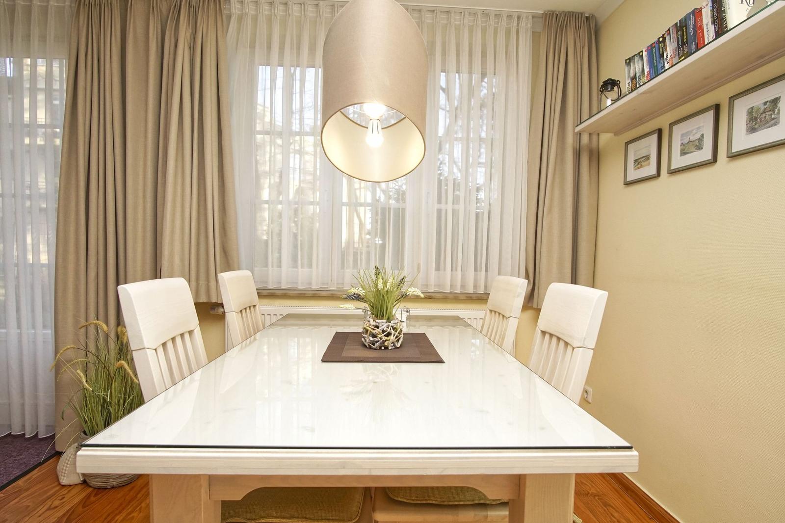 Esszimmer mit Tisch, Stühlen, Vorhängen und Deckenleuchte. Wand mit Bildern und Regal.