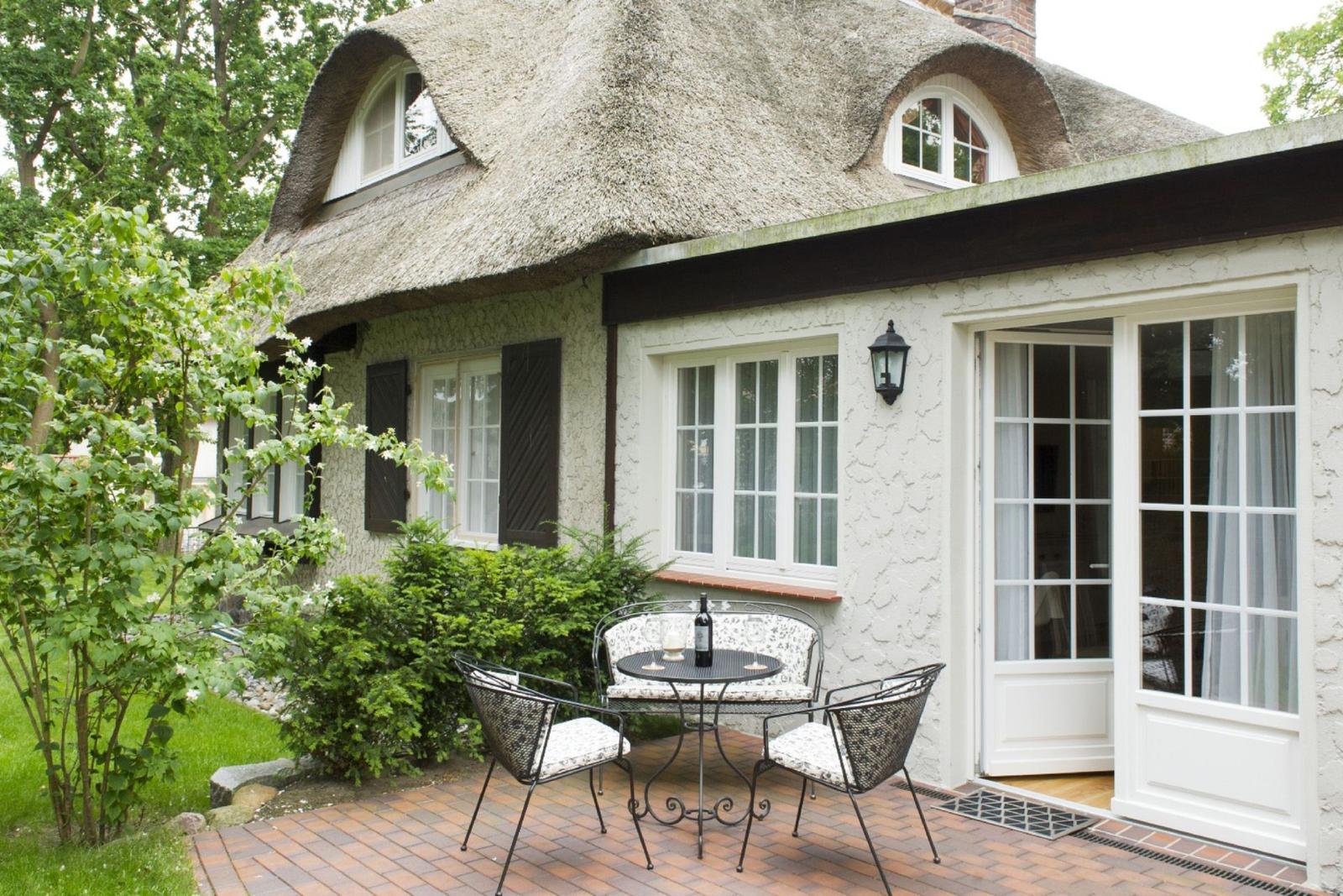 Haus mit Strohdach, Terrasse mit Tisch und Stühlen, Fenster und Glastüren.