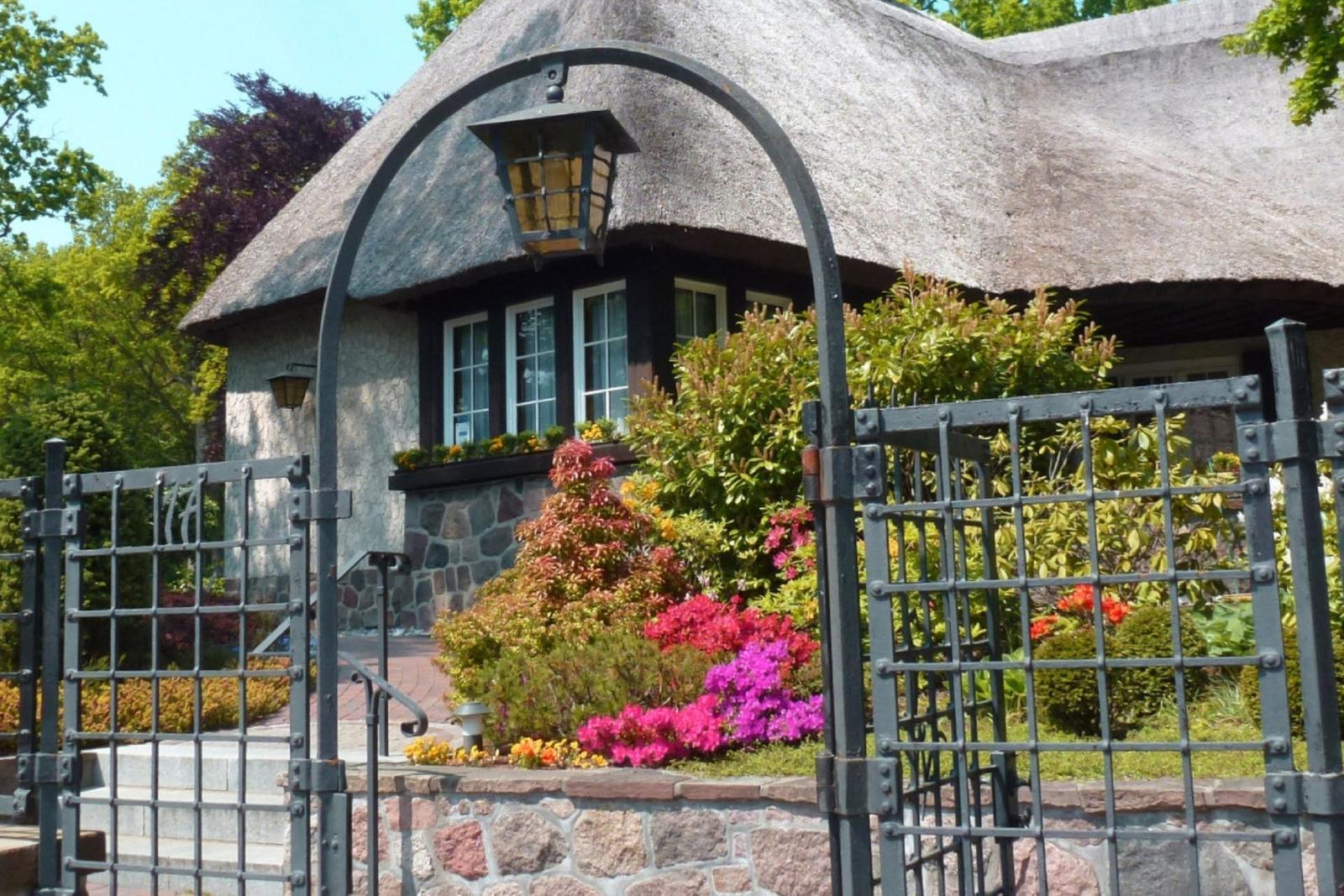 Haus mit Strohdach, Steinmauer und Metalltor. Blumenbeete und Fenster sind sichtbar.
