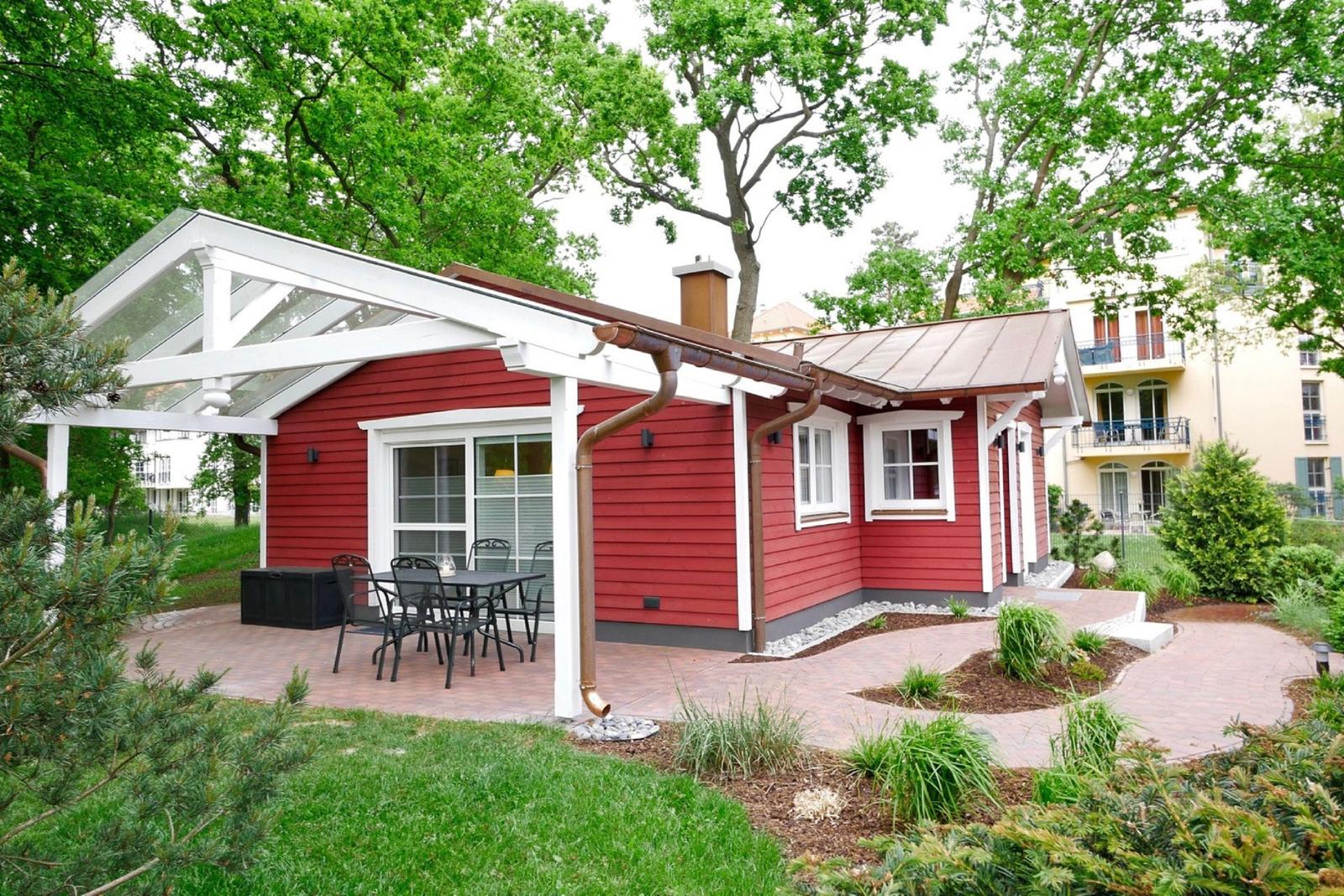 Rotes Holzhaus mit Terrasse und Sitzgelegenheit unter Glasdach. Umgeben von Grün und Bäumen.