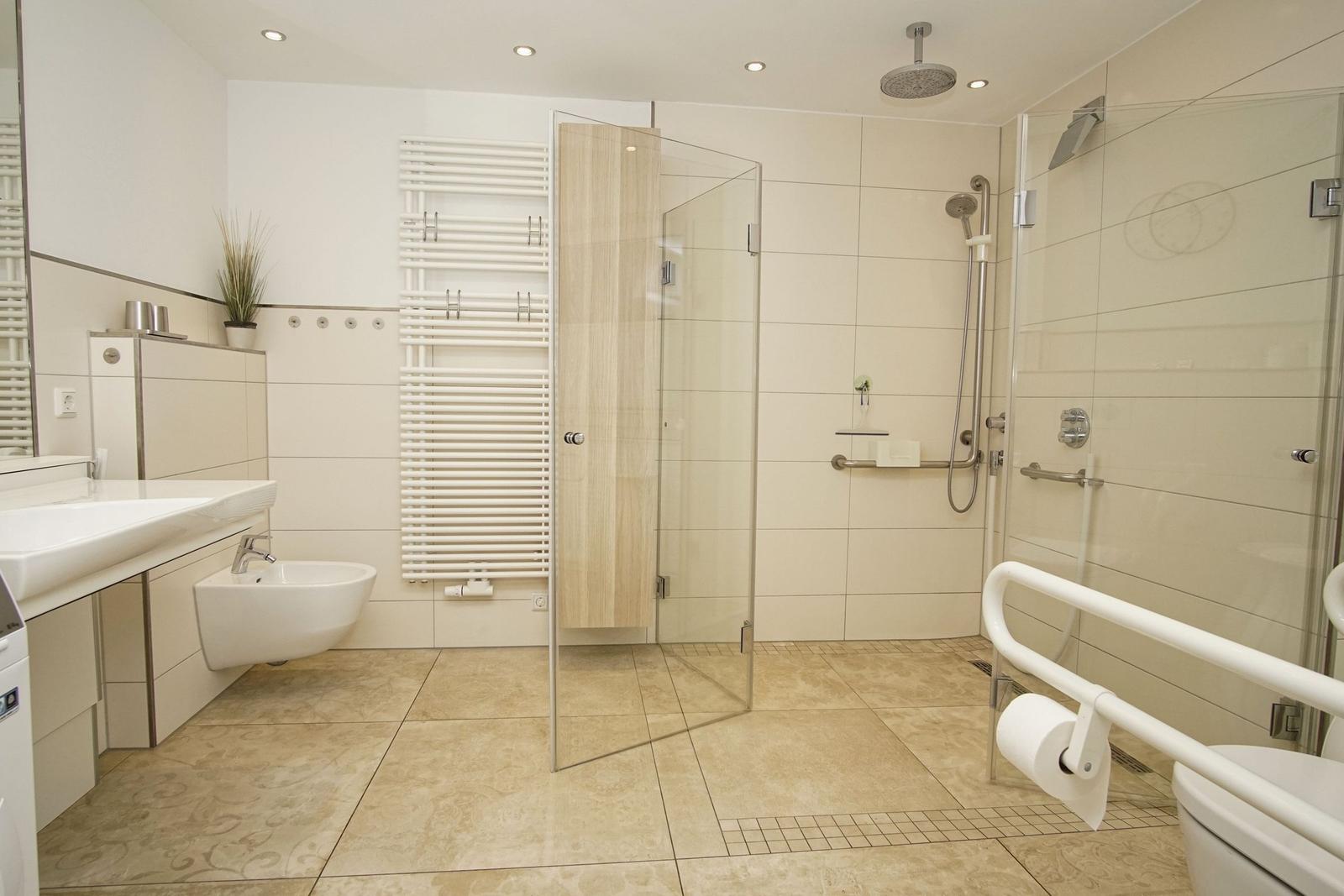 Badezimmer mit Dusche, WC und Waschbecken. Fliesenboden und Handlauf für barrierefreien Zugang.