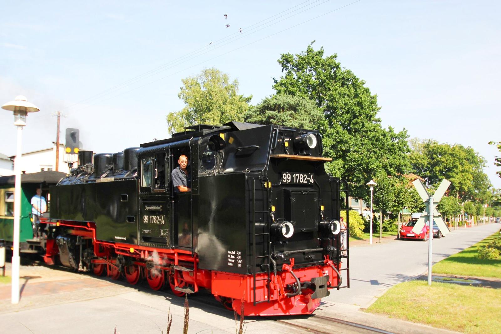 Dampflokomotive mit rotem Unterbau fährt durch eine städtische Umgebung mit Bäumen und Straßen.