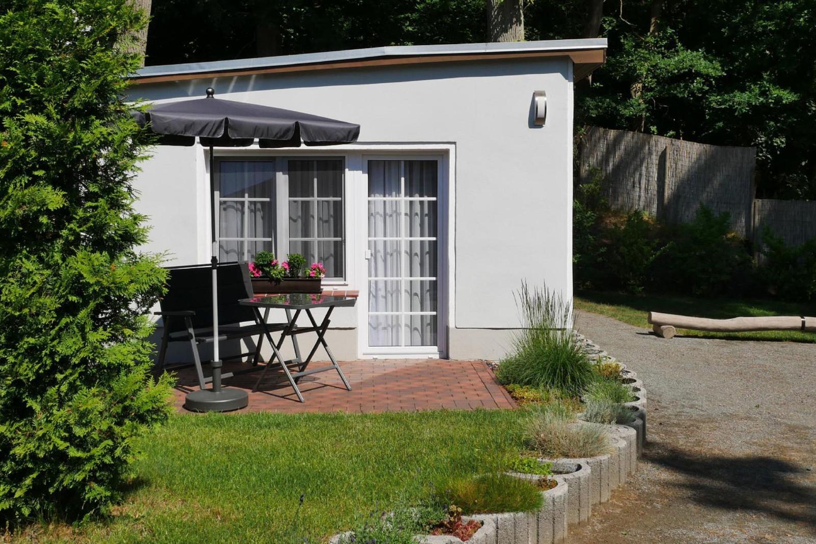 Wohnung mit Terrasse, Sonnenschutz, Tisch und Stühle. Grünanlage und gepflasterte Fläche.