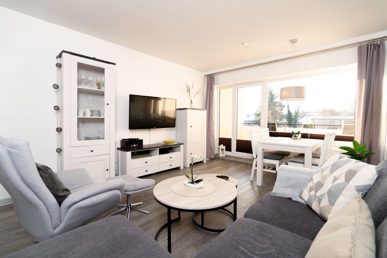 Wohn- und Essbereich mit Sofa, Sessel, Esstisch und TV. Balkon mit Sicht auf Grün.