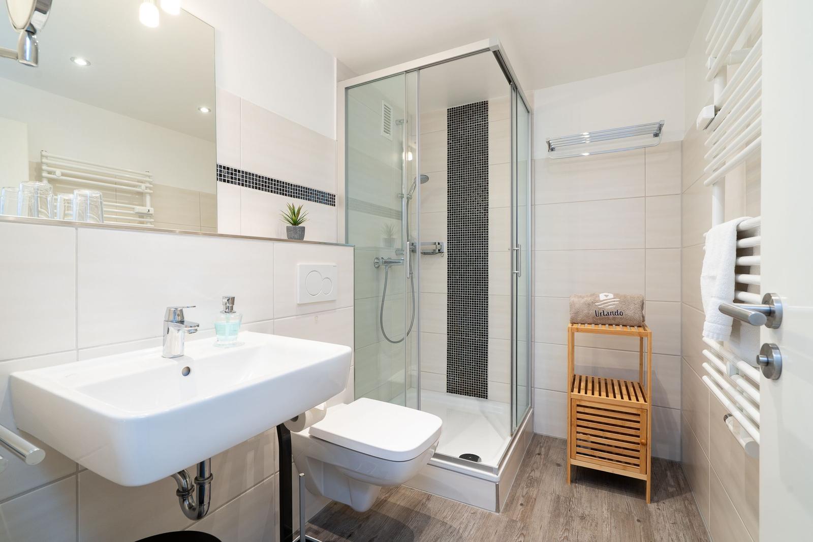 Badezimmer mit Dusche, Waschbecken und Toilette. Holzregal und Heizkörper.