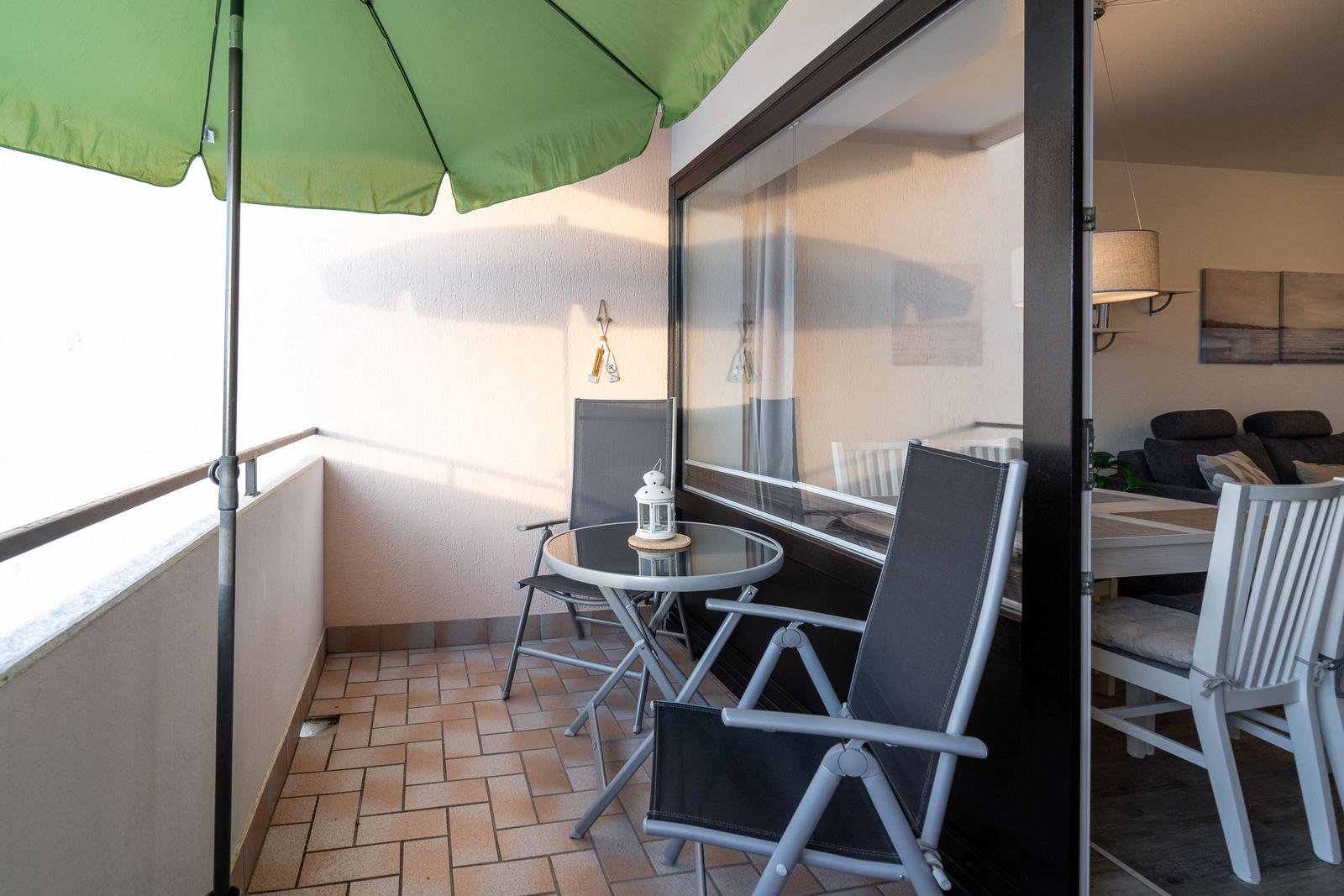 Terrasse mit grünem Sonnenschirm, Tisch und zwei Stühlen. Durchgang in Wohnbereich mit Sofa und Tisch.