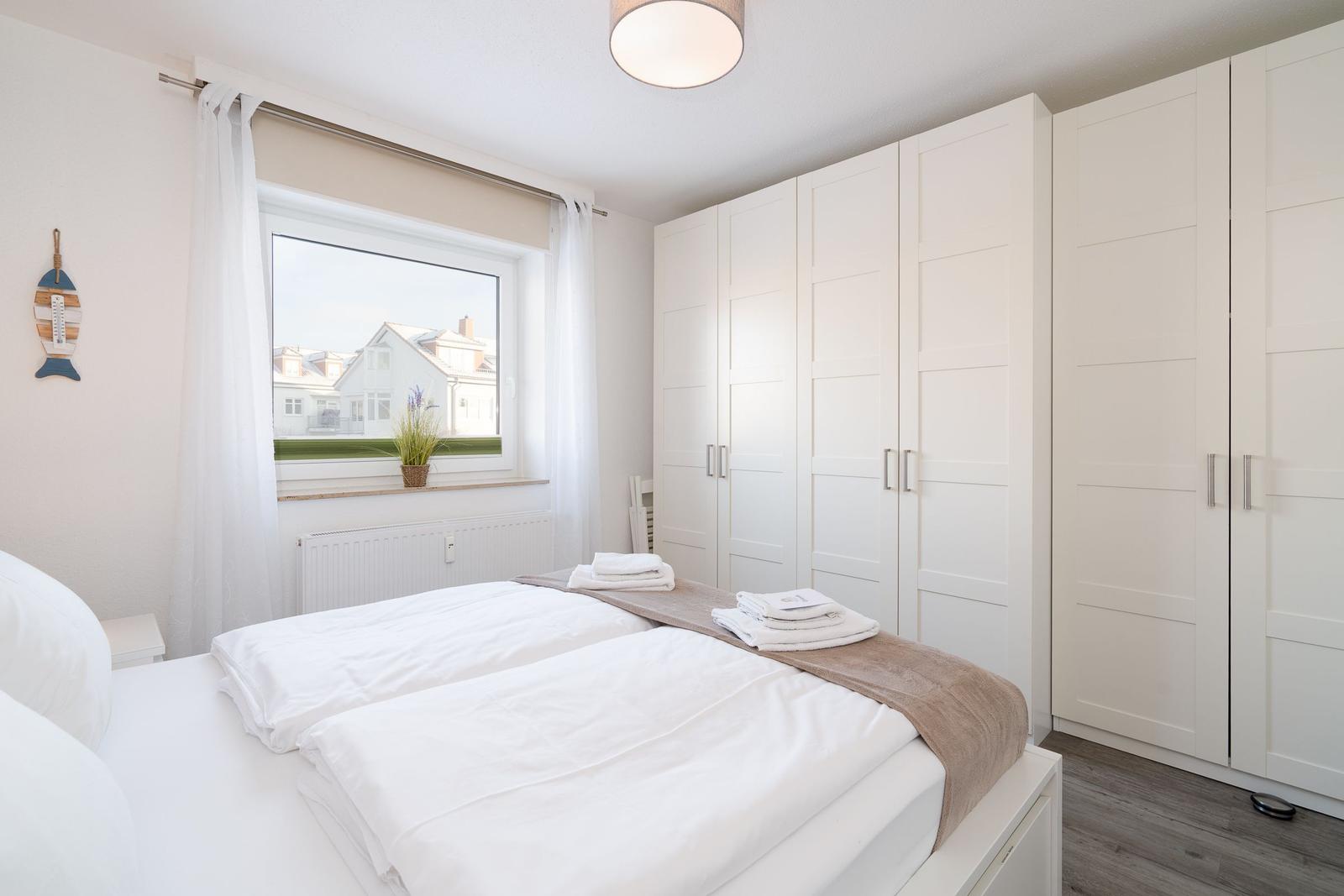 Schlafzimmer mit Doppelbett, weißen Schränken und Fensterblick auf Wohnhaus.