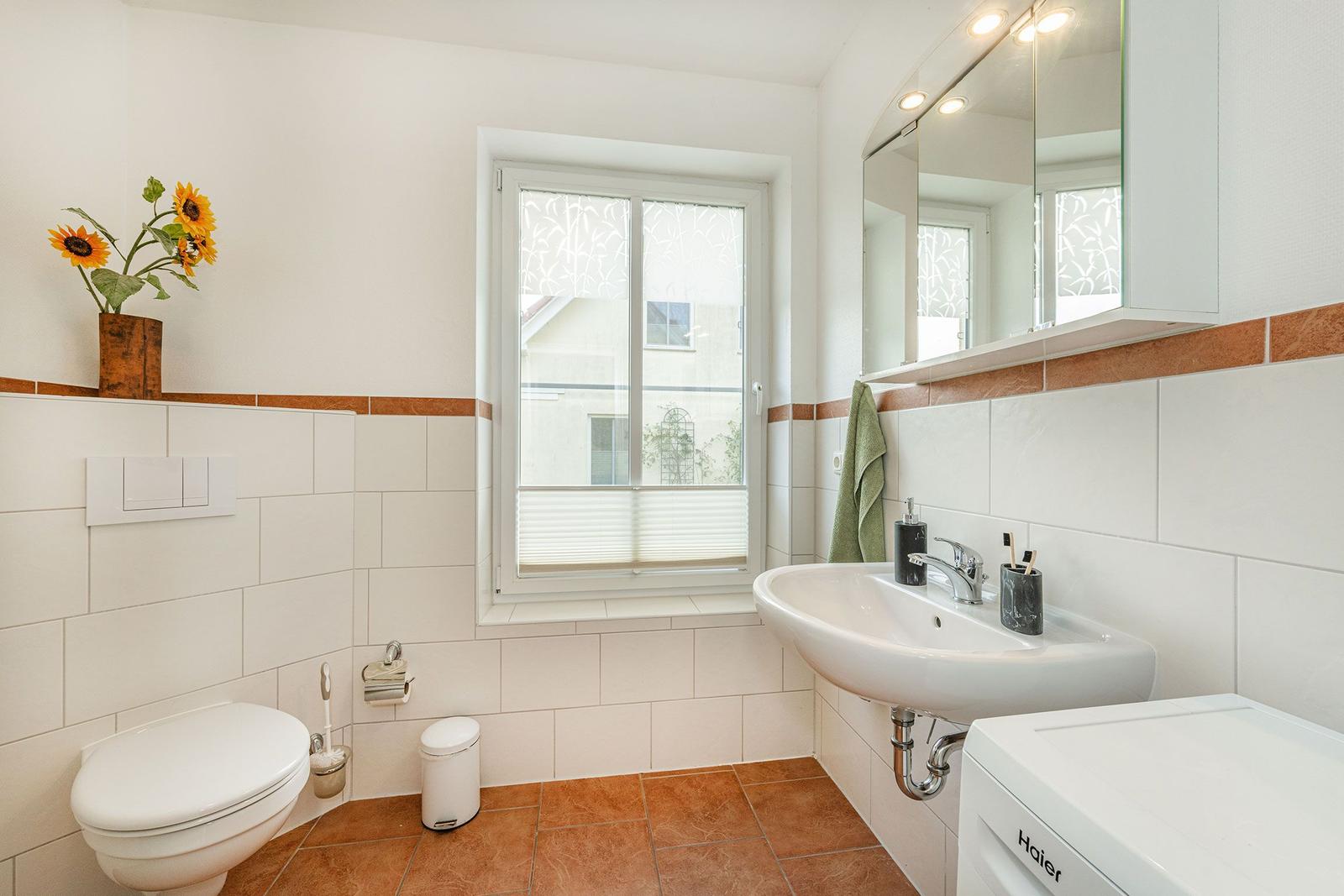 Badezimmer mit Toilette, Waschbecken und Fenster. Wand- und Bodenfliesen in Weiß und Terrakotta.