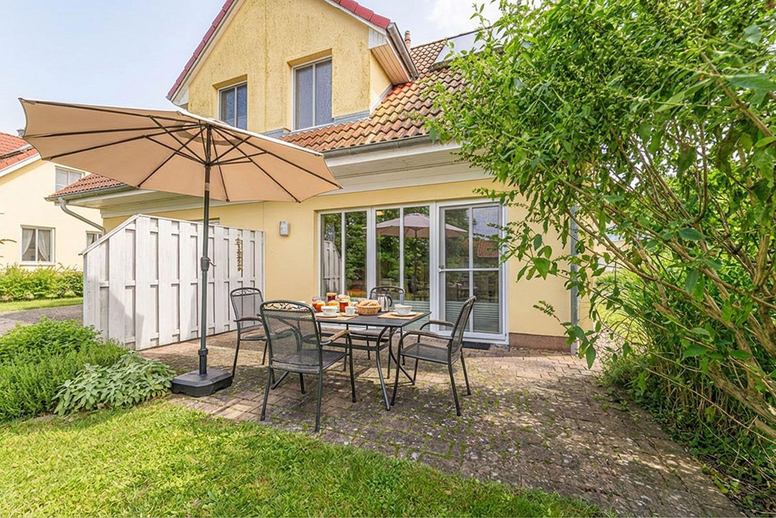 Terrasse mit Tisch, Stühlen und Sonnenschirm vor gelbem Haus mit Glasfront.