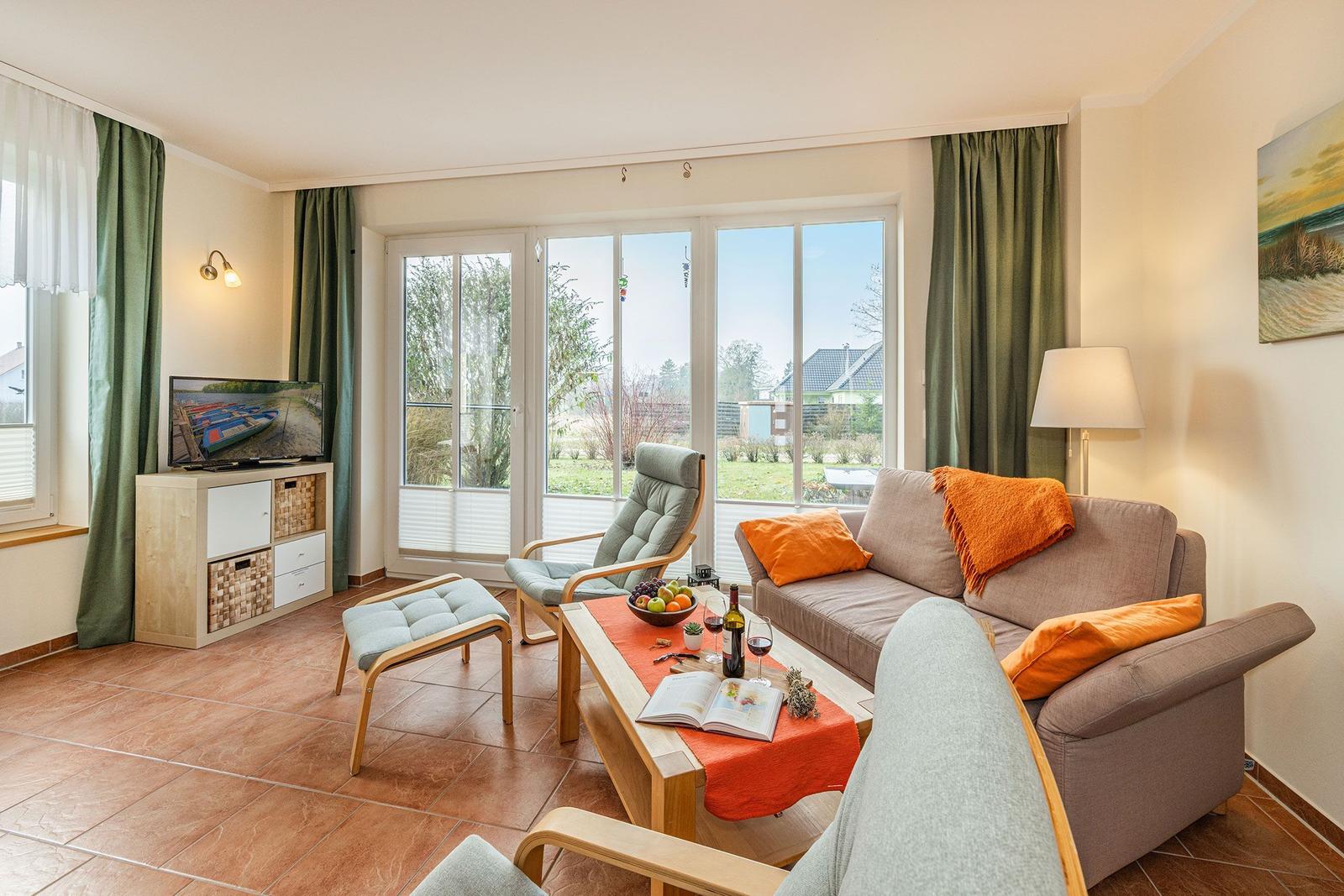 Wohnzimmer mit Sofa, Sessel und Fernseher. Große Glastüren führen auf die Terrasse.