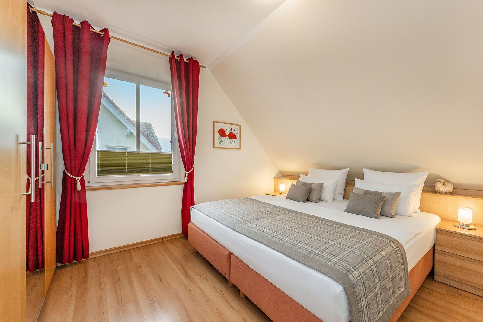 Schlafzimmer mit Doppelbett, roten Vorhängen und Holzboden. Fenster mit Blick auf Dach.