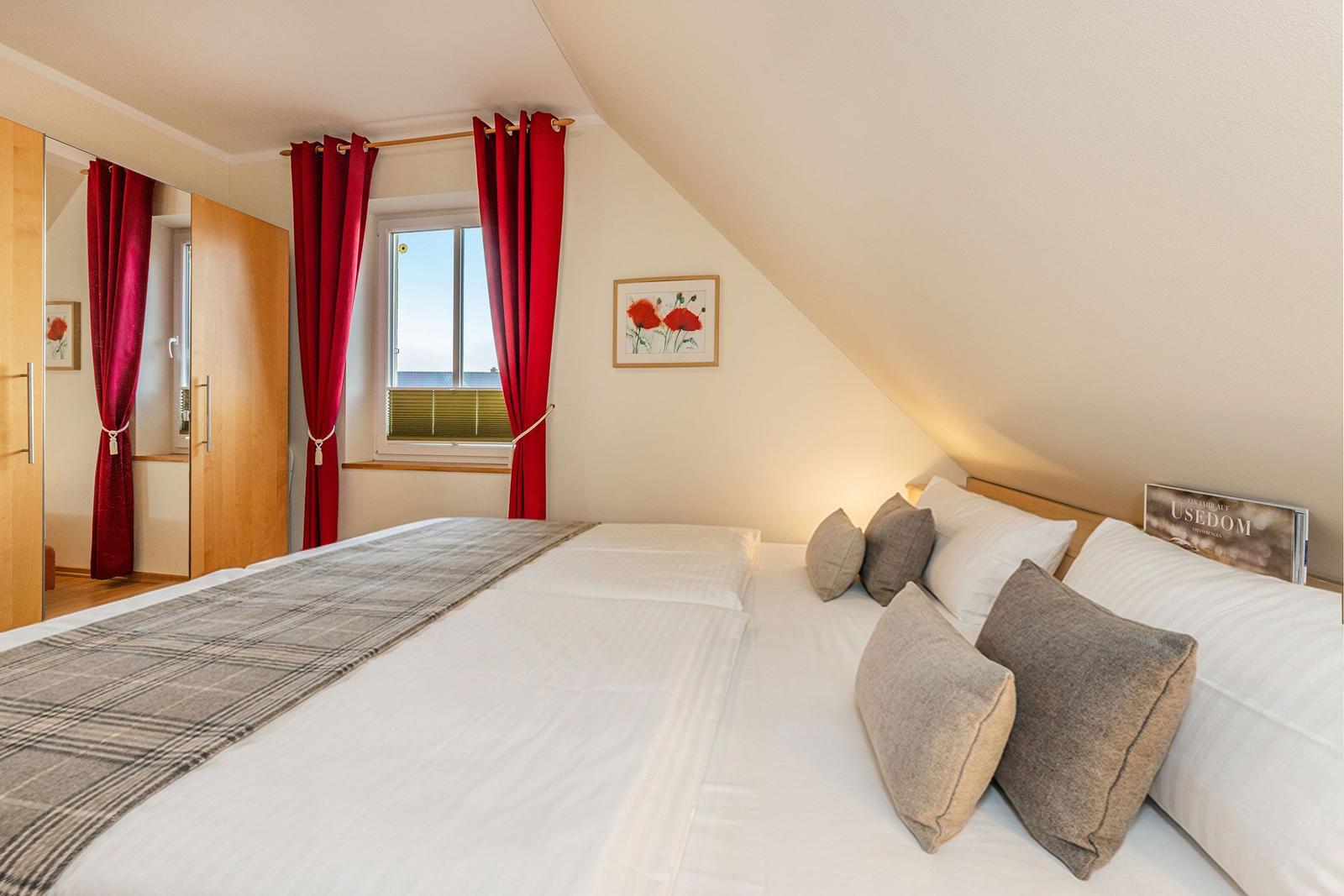 Schlafzimmer mit Dachschrägen, Bett mit Kissen, rote Vorhänge und Fenster.