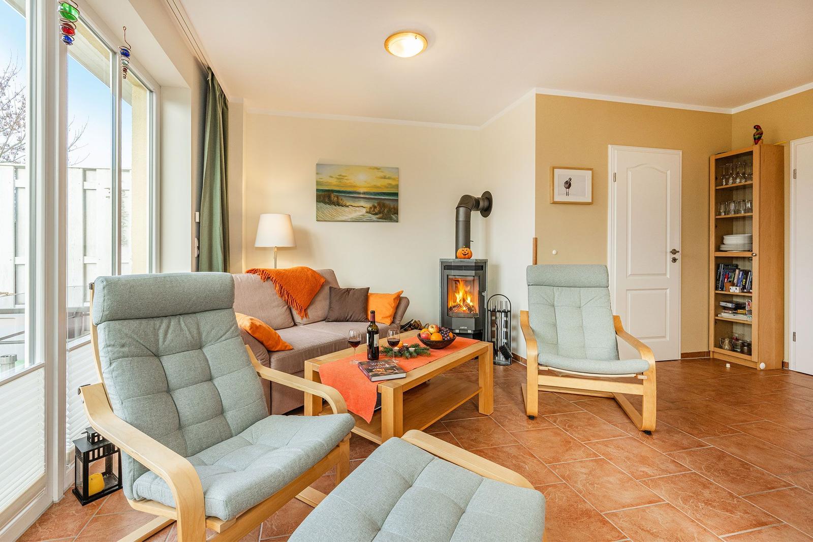 Wohnzimmer mit Kamin, Sofa, zwei Sessel und Holztisch. Große Fenster und Bücherregal.