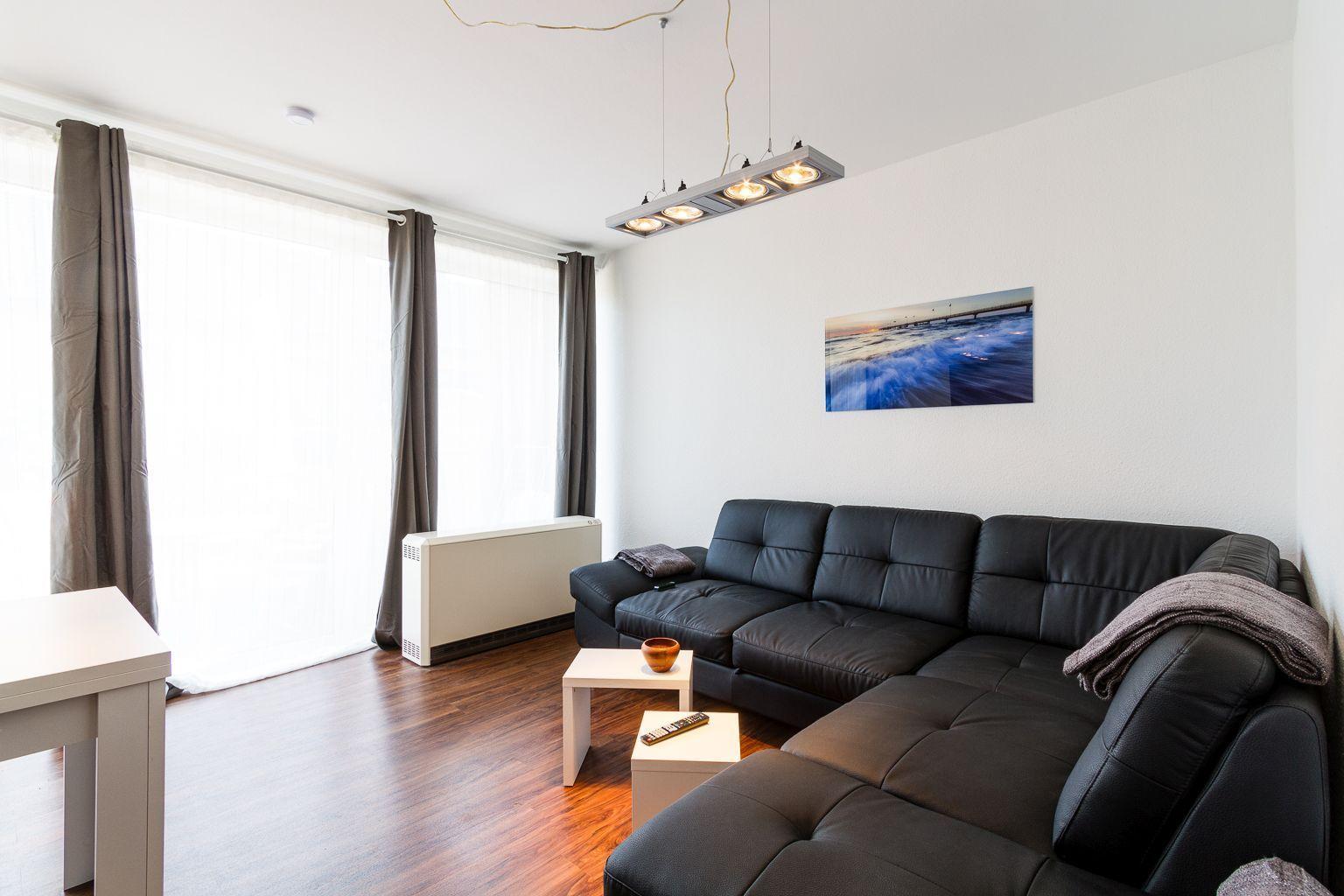 Wohnzimmer mit schwarzer Ledercouch, weißen Tischen und Holzboden. An der Wand hängt ein Bild.