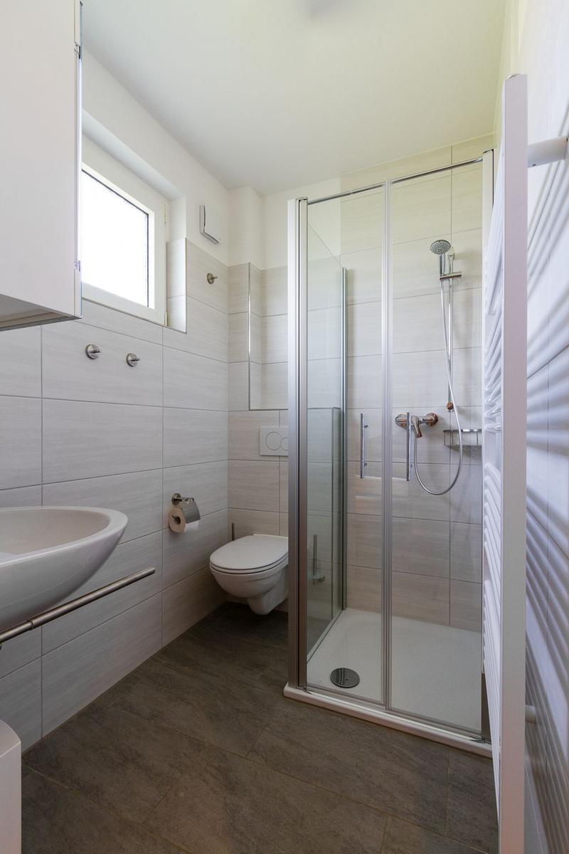Badezimmer mit Dusche, Toilette und Waschbecken. Fliesenwand und Fenster sichtbar.