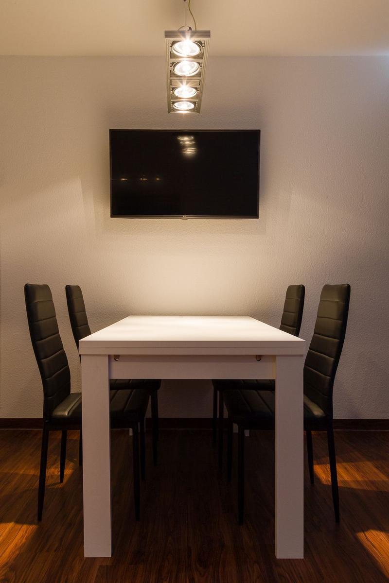 Esszimmer mit weißem Tisch, vier schwarzen Stühlen und Wand-TV unter einer Deckenleuchte.