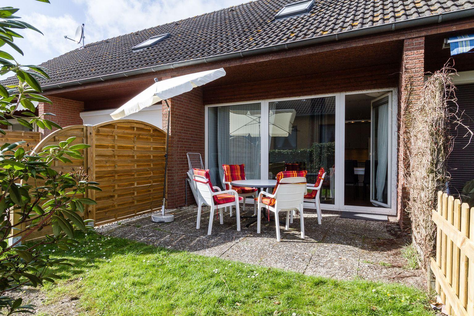 Terrasse mit Tisch, Stühlen und Sonnenschirm. Hintergrund: Haus mit Glasfront und Holzzaun.