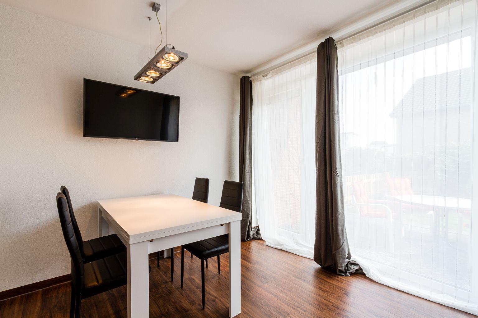 Esszimmer mit Tisch, Stühlen, Fernseher und großem Fenster mit Blick auf Balkon.