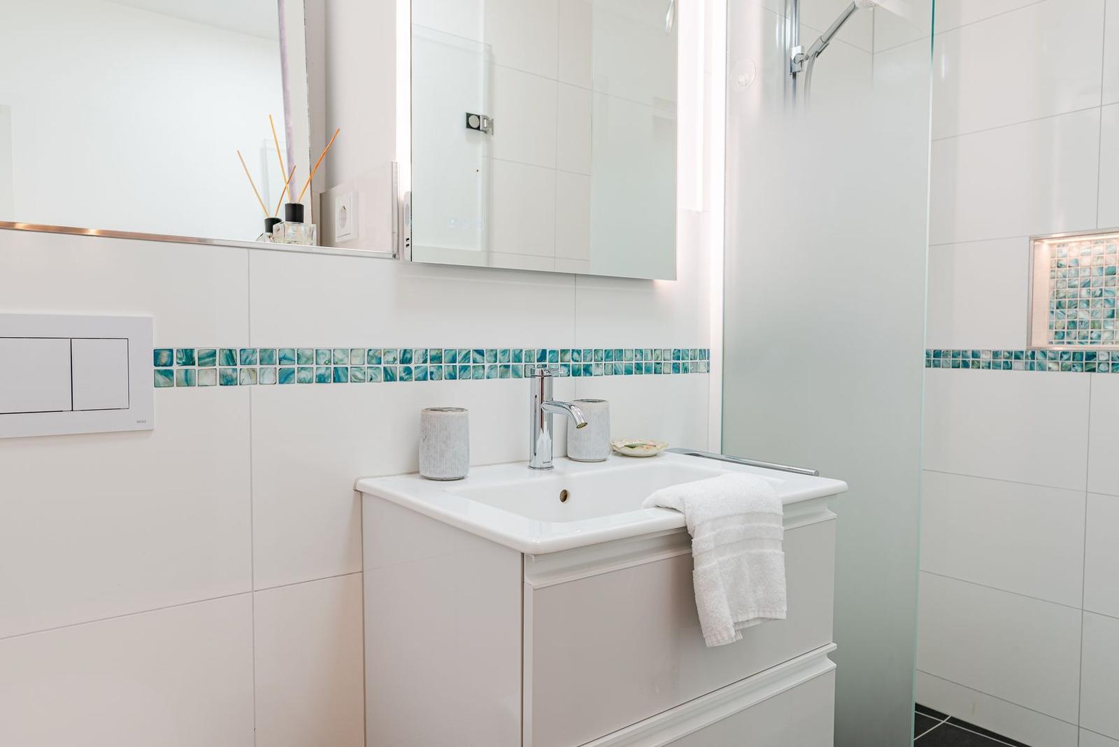 Bad mit Waschbecken, Spiegel und Dusche. Wandfliesen mit blauem Mosaikstreifen.