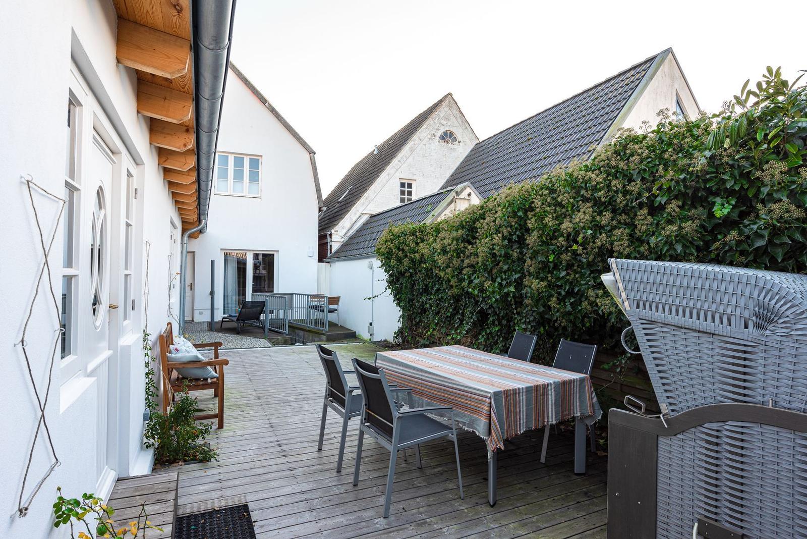 Hinterhof mit Holzterrasse, Tisch, Stühlen und Beerenhecke. Hintergrund: weiße Häuser mit Dächern.