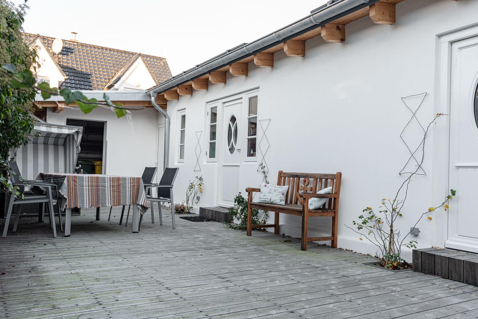 Terrasse mit Holzbank, Tisch und Stühlen. Weiße Wand, Fenster und Tür.