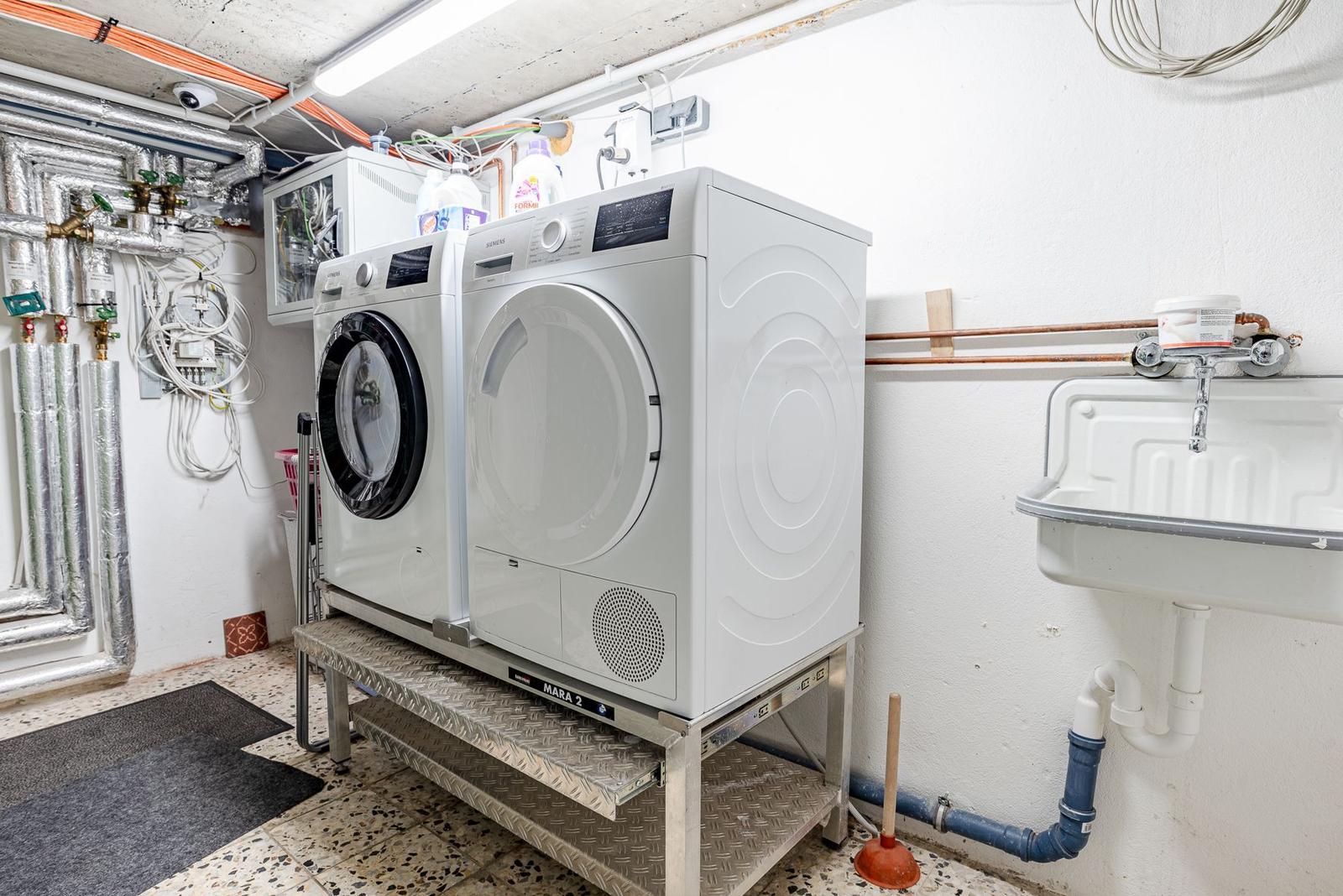 Wasch- und Trockner stehen auf Metallgestell. An der Wand hängt ein Waschbecken mit Armatur.