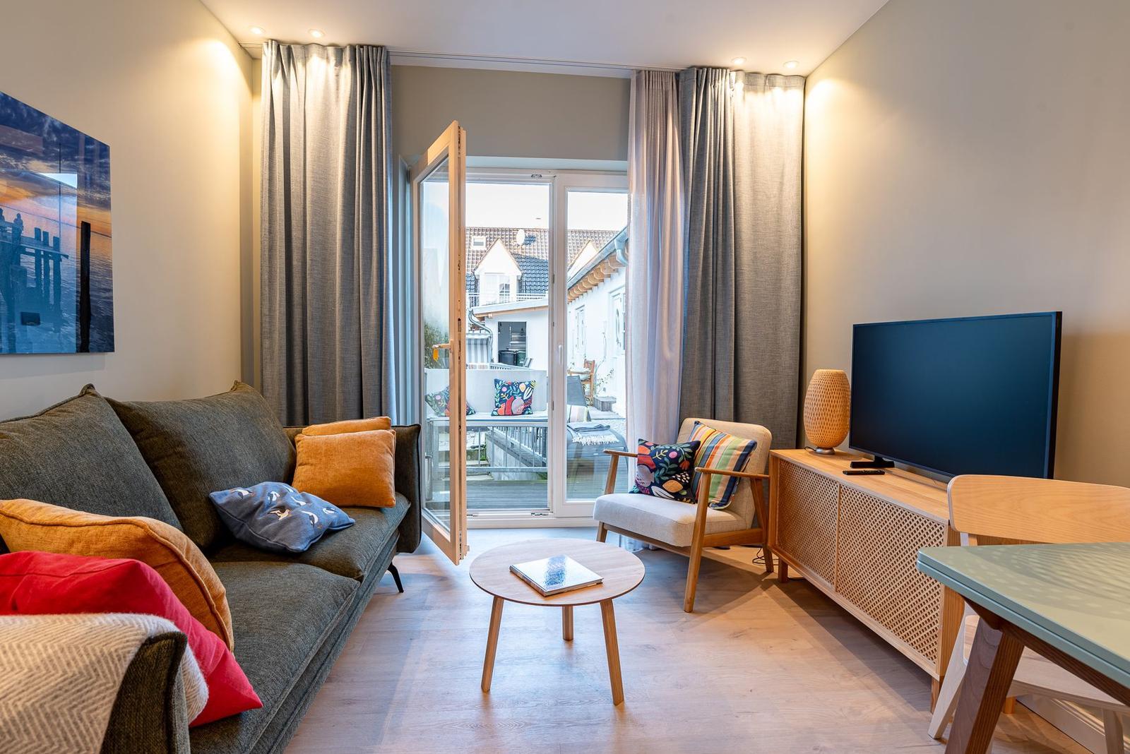 Wohnzimmer mit Sofa, Sessel und Fernseher. Durchgang zur Terrasse mit Blick auf Wohnhäuser.