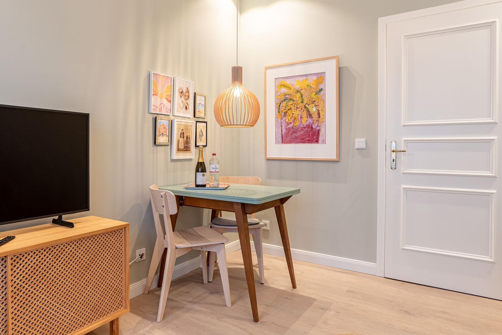 Essbereich mit Tisch, Stühlen und Wandbildern. Fernseher auf Holzregal.