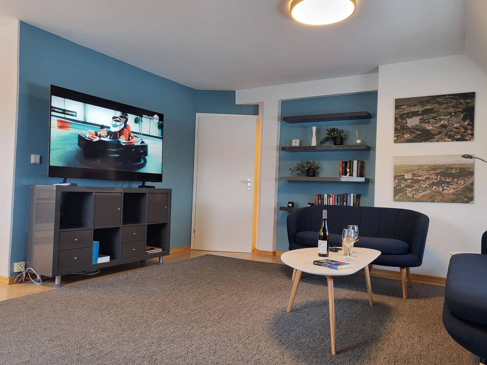 Wohnzimmer mit Fernseher, Sofa und Tisch. Wand mit Regalen und Bildern.