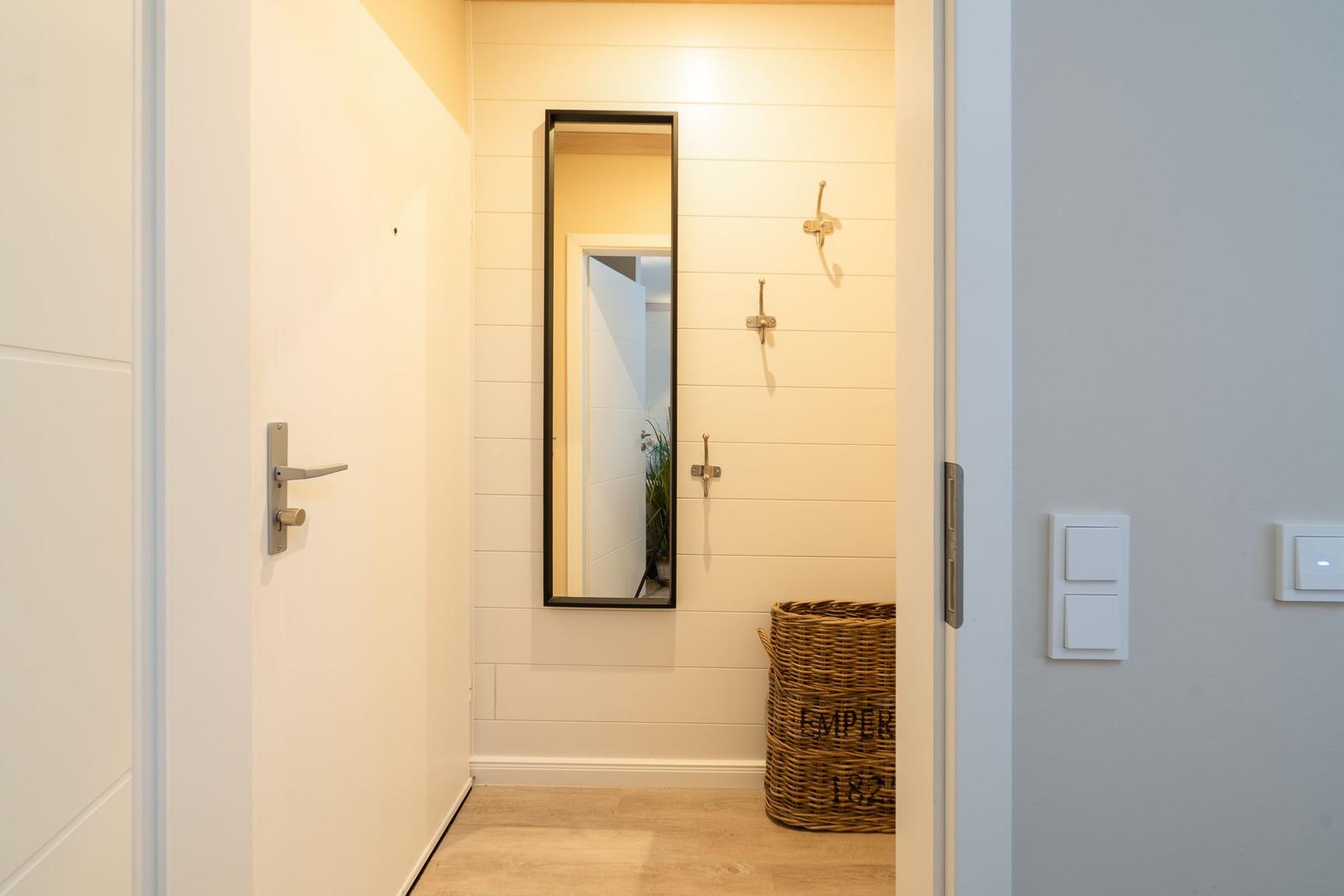 Eingangsbereich mit Spiegel, Haken und Korb. Holzdielen und weiße Wände.