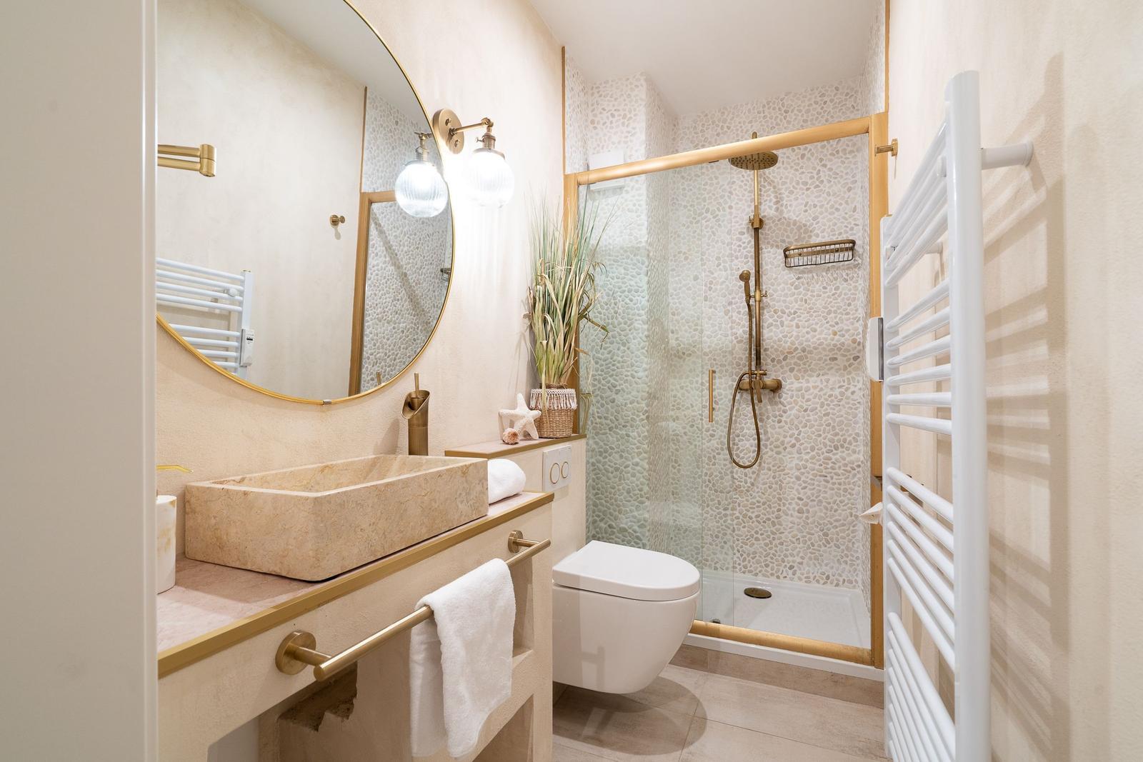 Badezimmer mit Dusche, Waschtisch und Toilette. Holzrahmen und goldfarbene Armaturen.