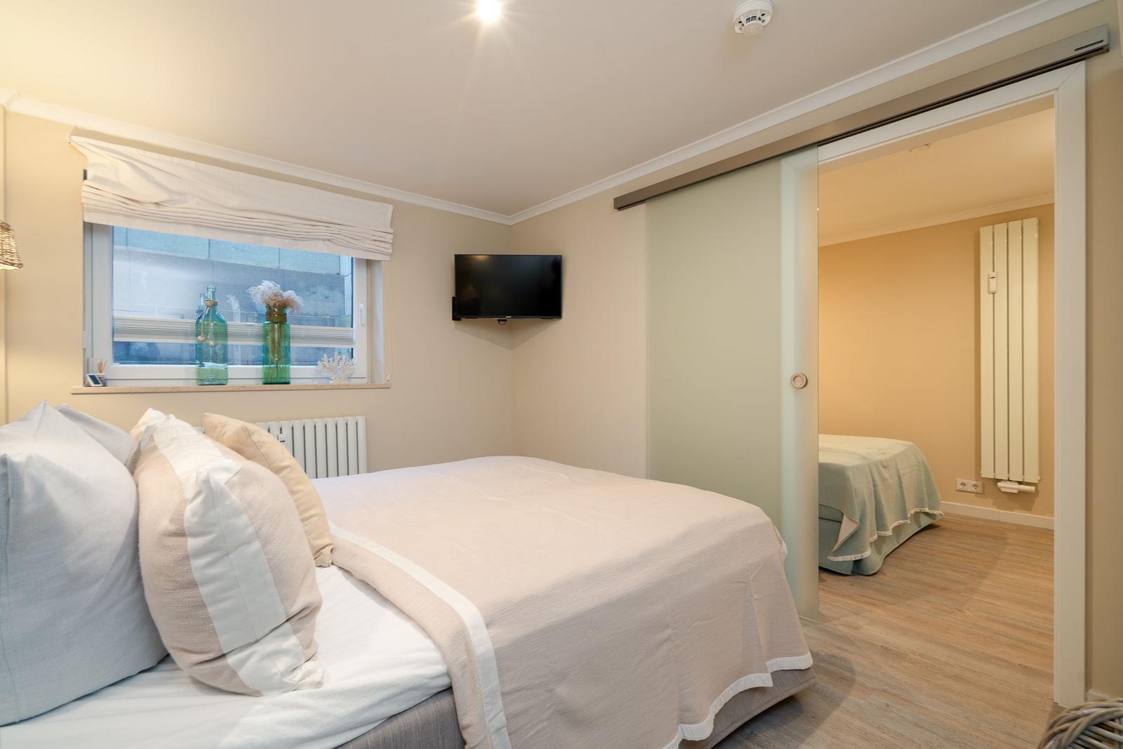 Schlafzimmer mit Bett, TV und Fenster. Durch Glastür ist zweites Schlafzimmer sichtbar.